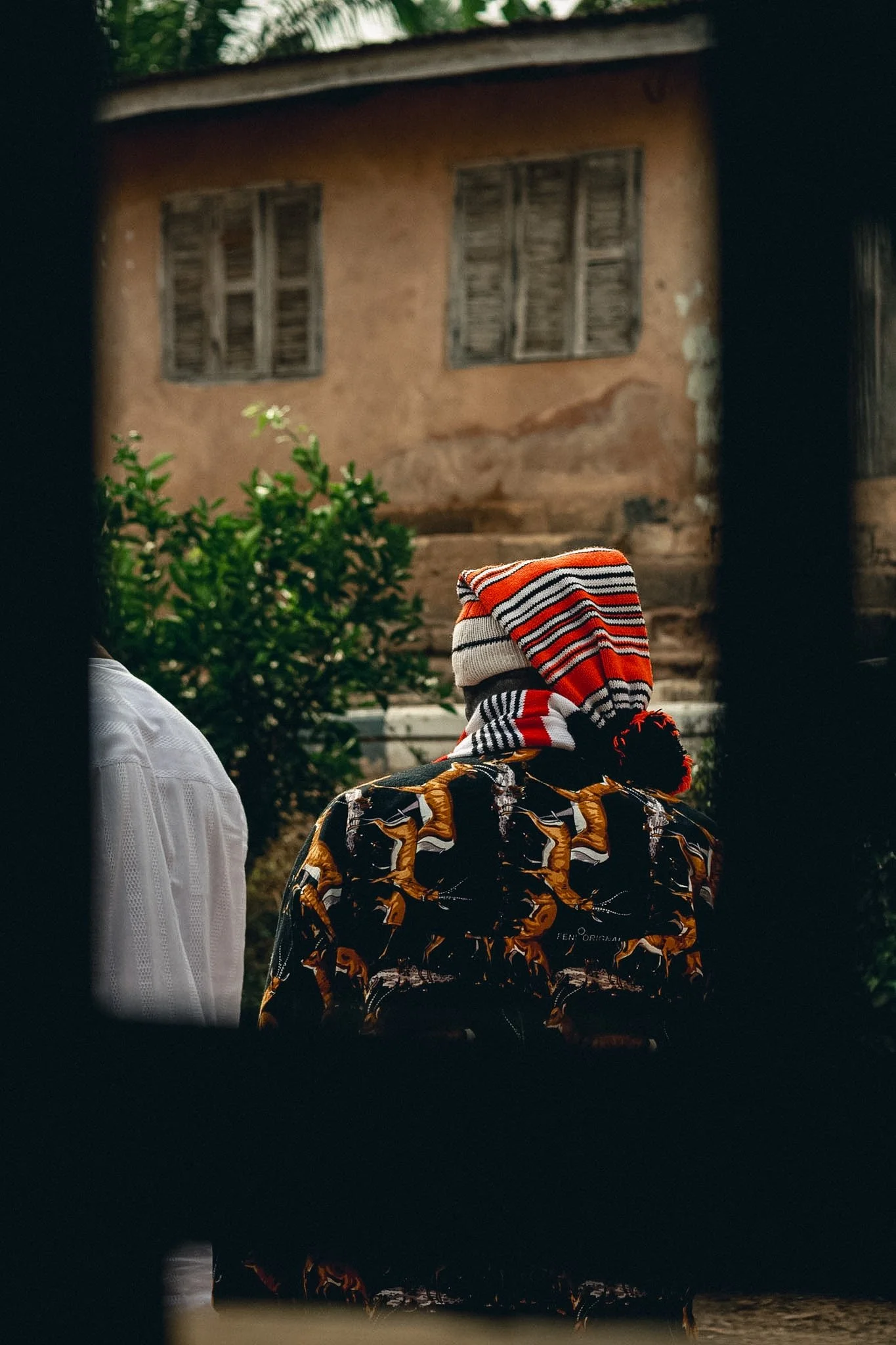 View of a mud house with two closed wooden windows, partially obscured by dark framing in the foreground, greenery in front of the house, and two people wearing patterned clothing and a striped hat facing the house.