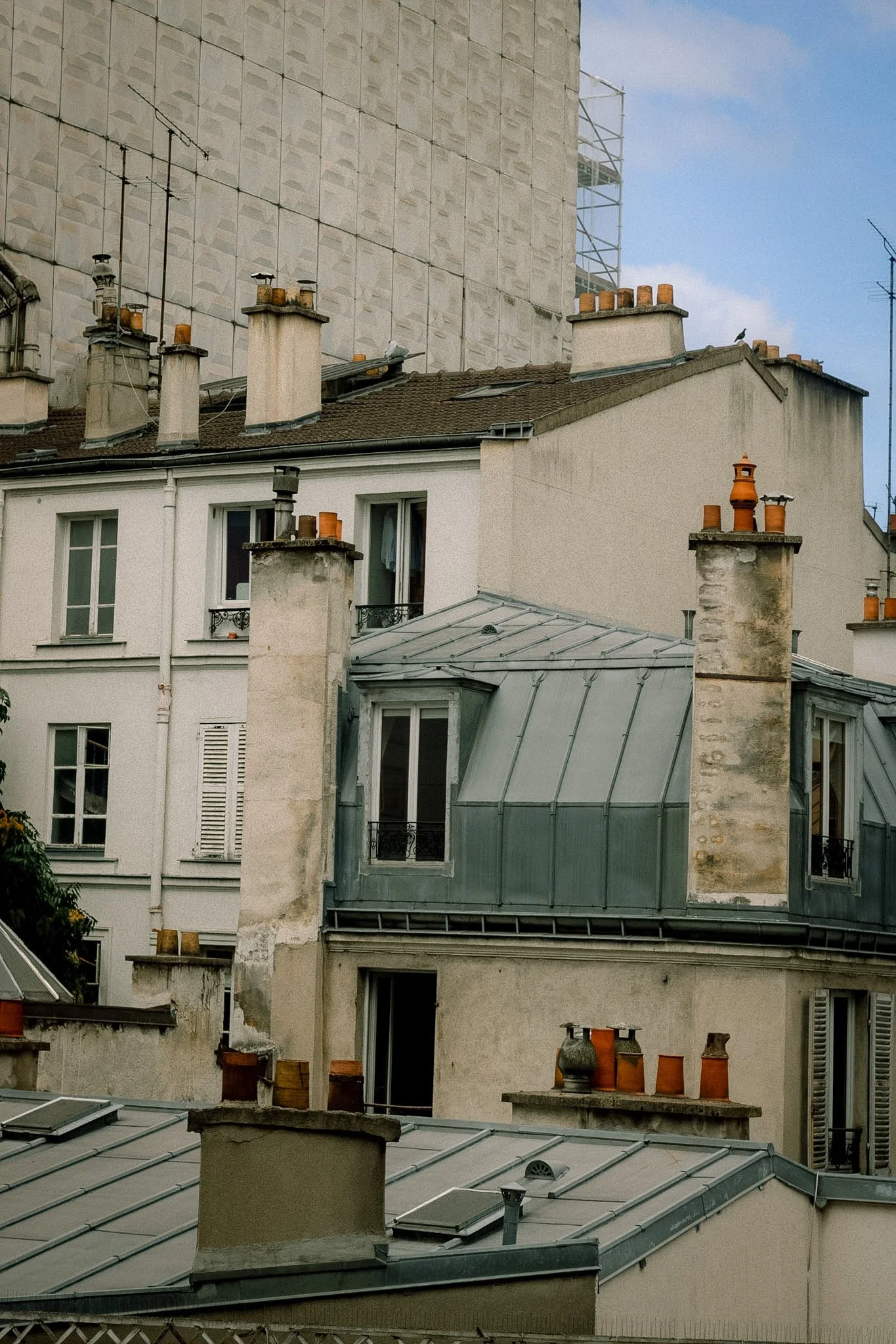 Rooftops of Paris apartment buildings with multiple chimneys and skylights, some with scaffolding in the background, under a partly cloudy sky.