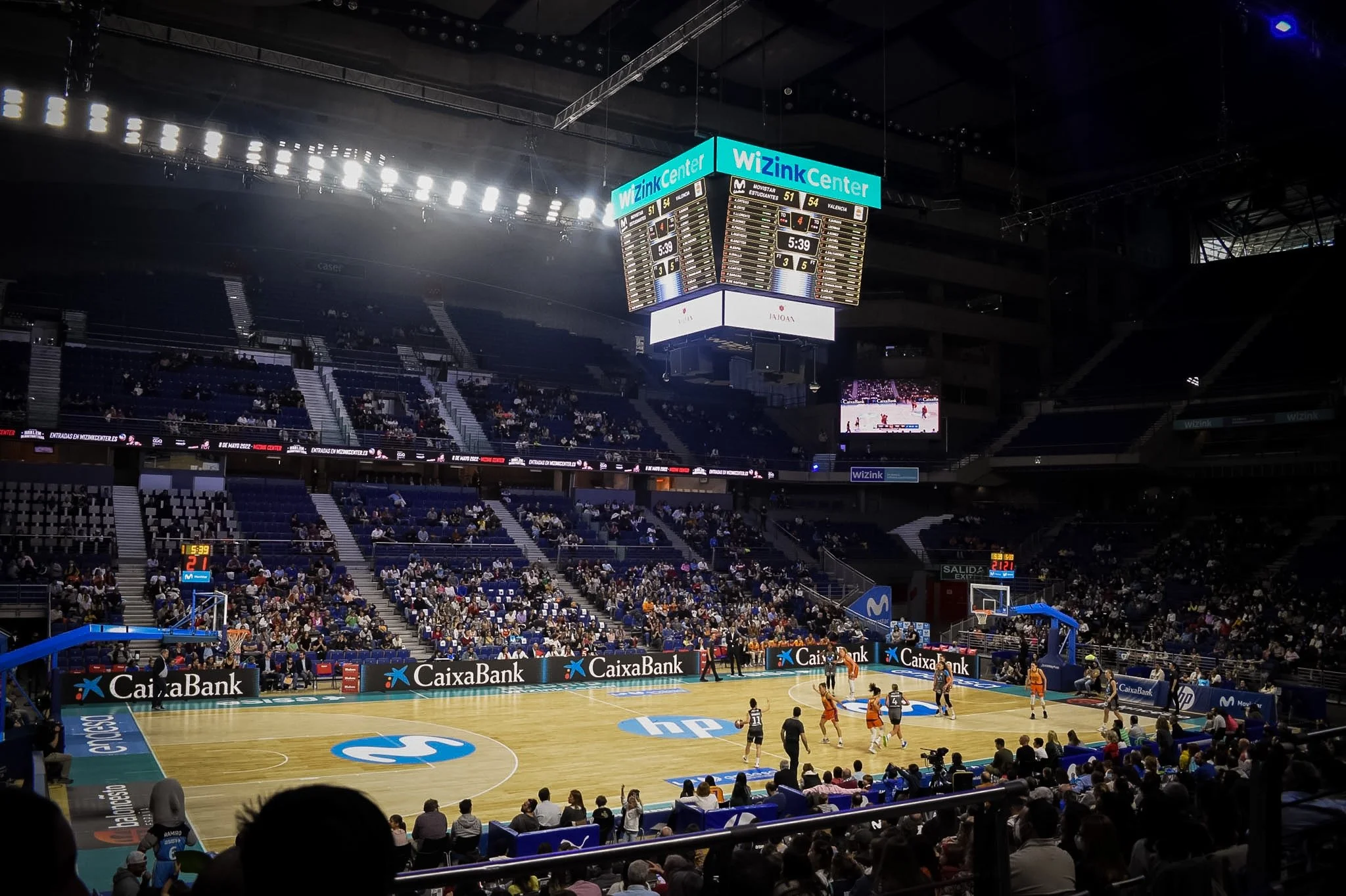 An indoor basketball arena with players on the court, audience in the stands, and scoreboard showing game details.