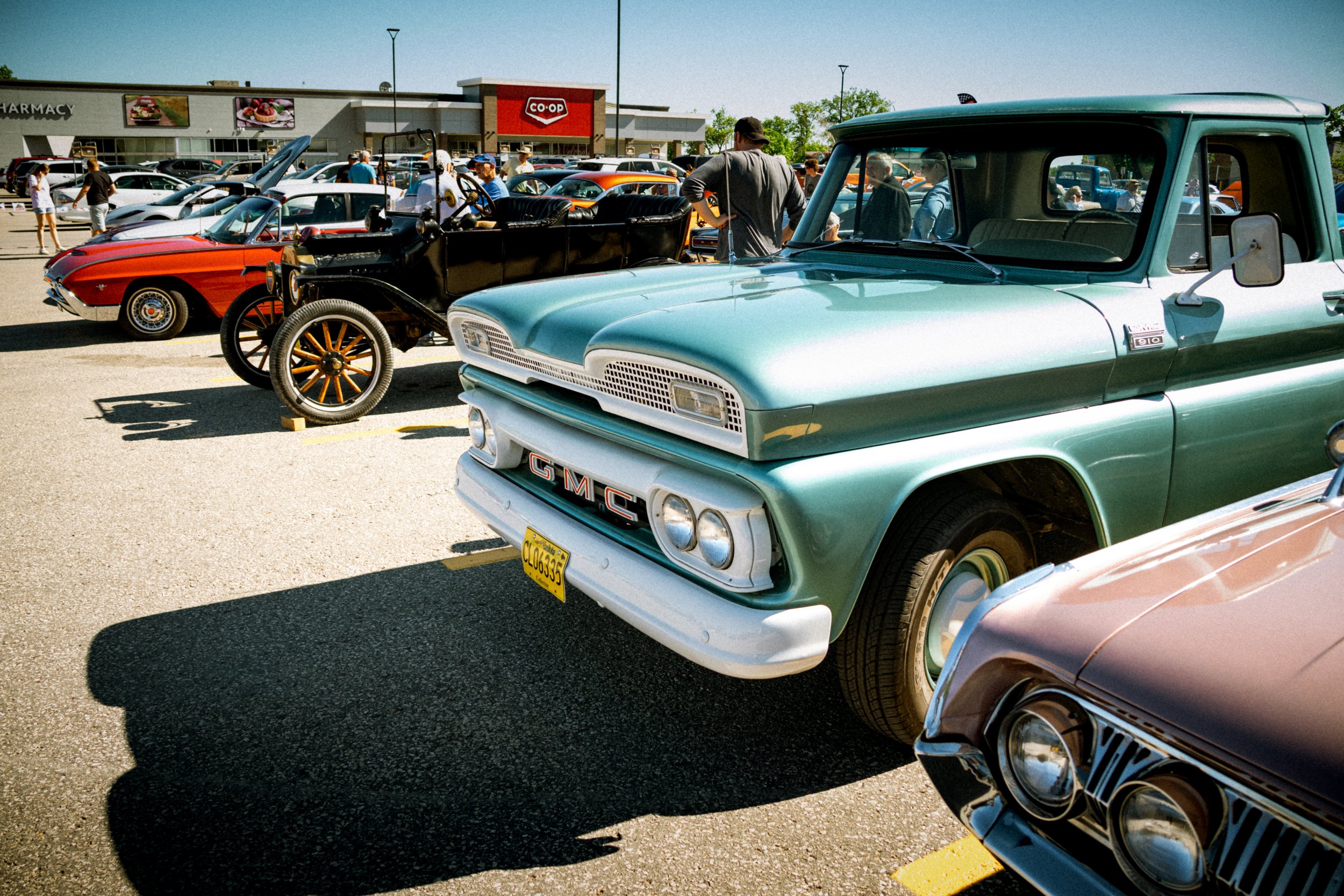 A parking lot filled with vintage cars and people attending a car show outside a shopping center with a Co-op store. The sky is clear and sunny.
