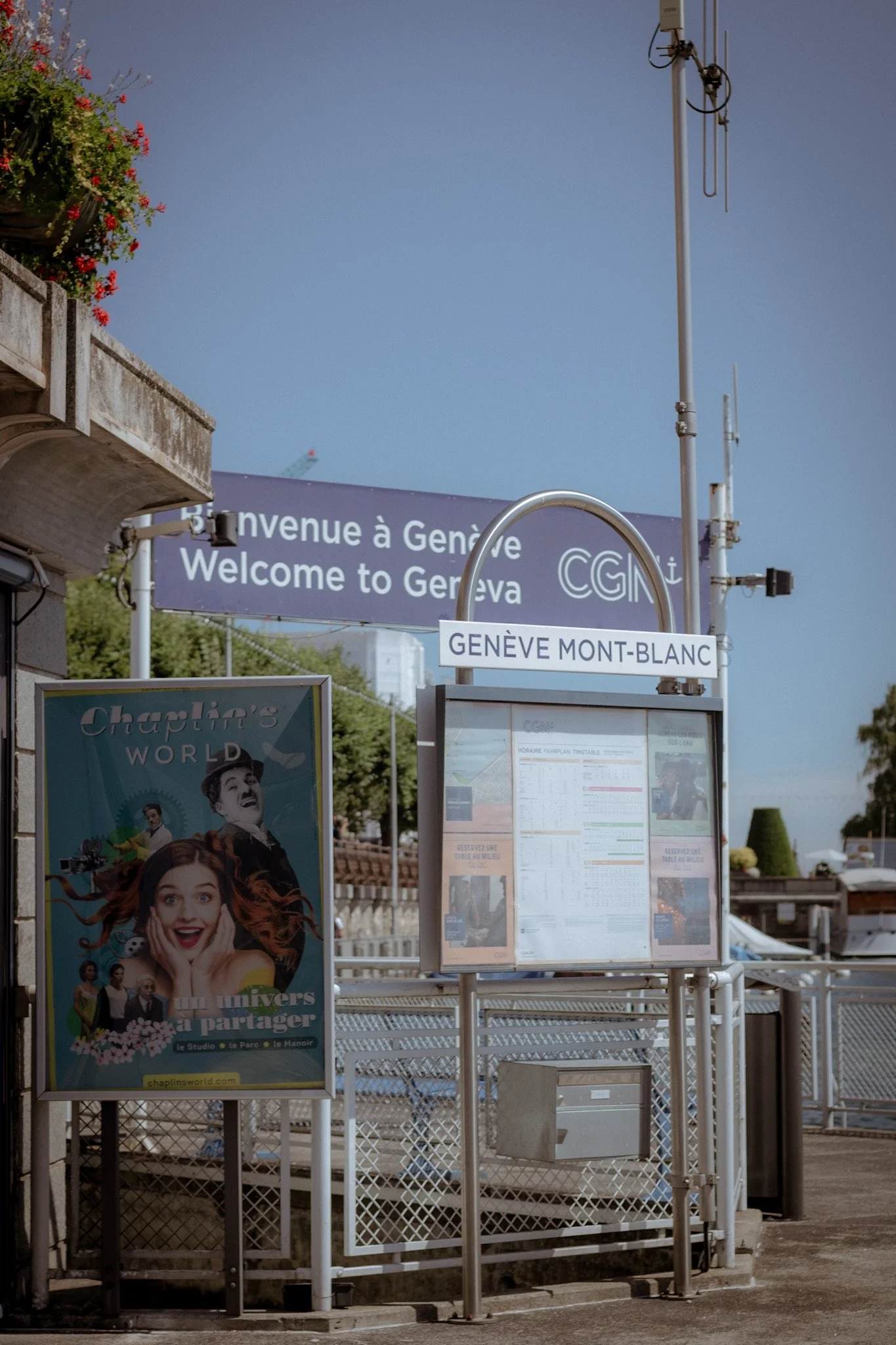 A public transportation bus stop in Geneva Mont-Blanc, France, with signs and posters, including a colorful advertisement featuring smiling people and a poster with schedules, against a clear blue sky.