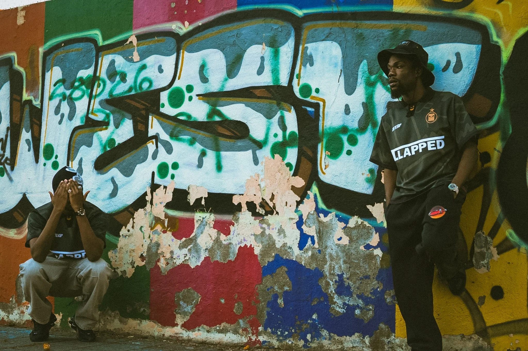 Three people in front of a colorful graffiti wall. One person is standing with hands covering their face, another is standing with arms crossed, and the third person is looking to the right, wearing a bucket hat and a black shirt with the word "LAPPE