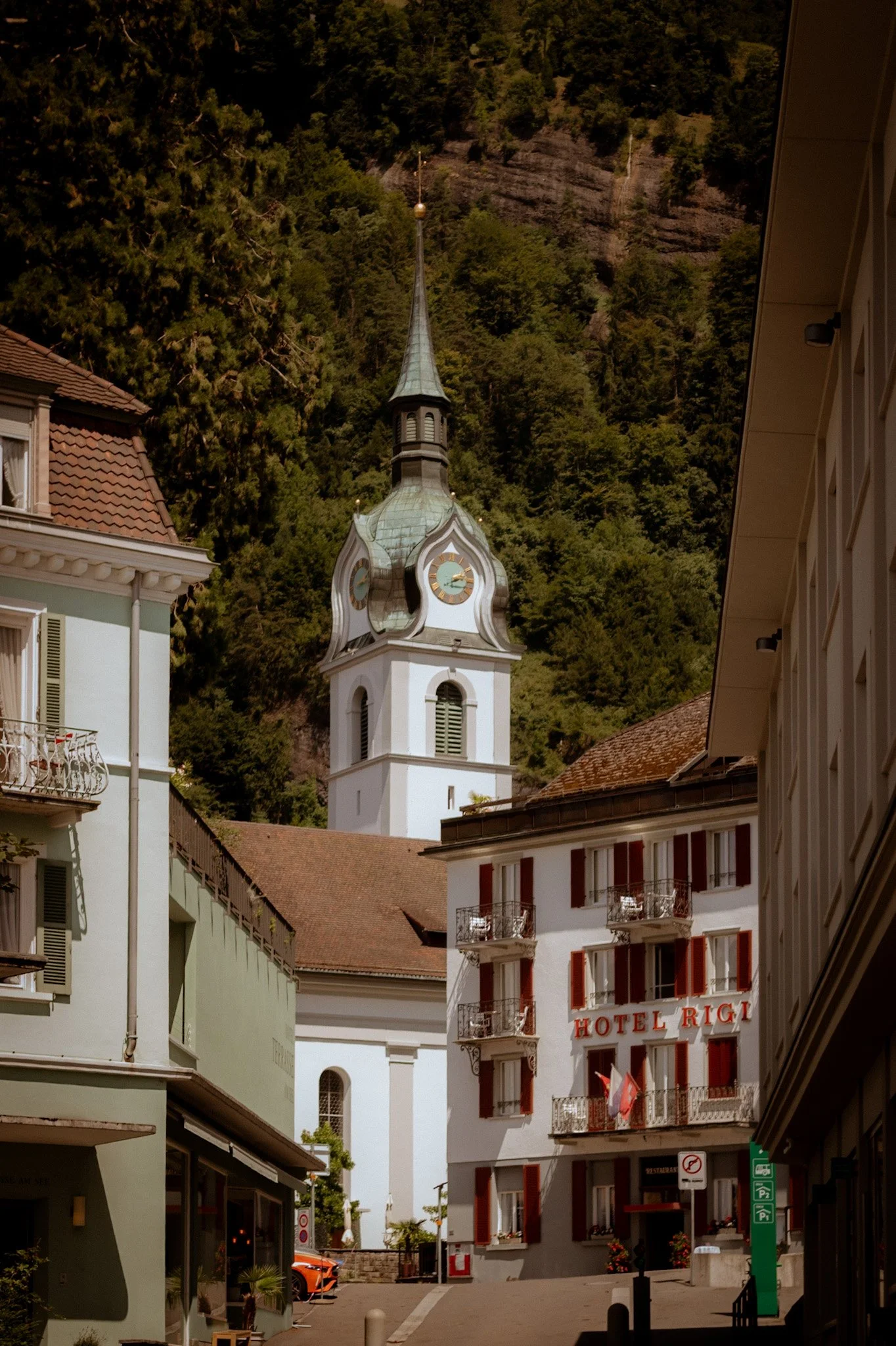 A street scene featuring a white church with a tall steeple and clock faces, surrounded by buildings including a hotel named Hotel Rigi with balconies. There are parked cars, signs, and greenery in the background, with a mountain covered in trees beh