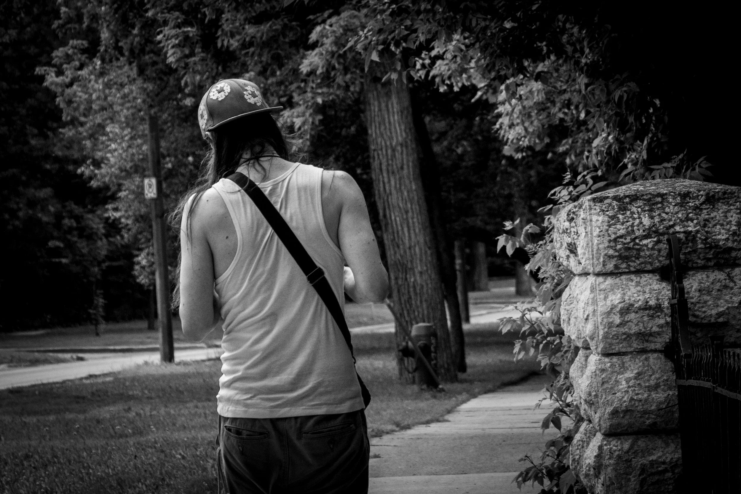 A woman walking outdoors wearing a tank top and a hat with flowers, with a strap across her chest, near a stone wall and trees.