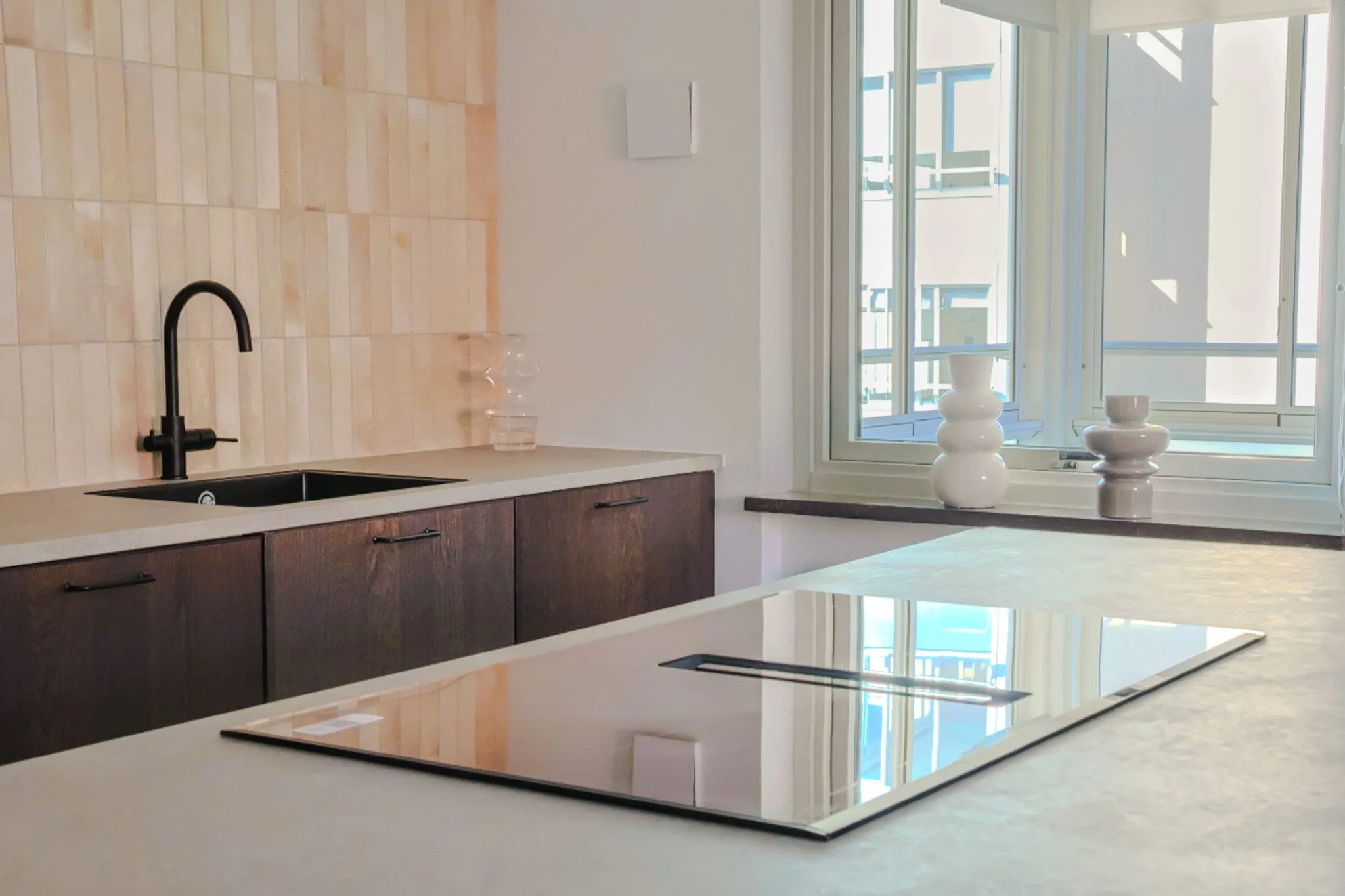 Modern kitchen with wooden cabinets, black faucet, beige tiled backsplash, white countertop, and a large window with decorative white vases on the sill.