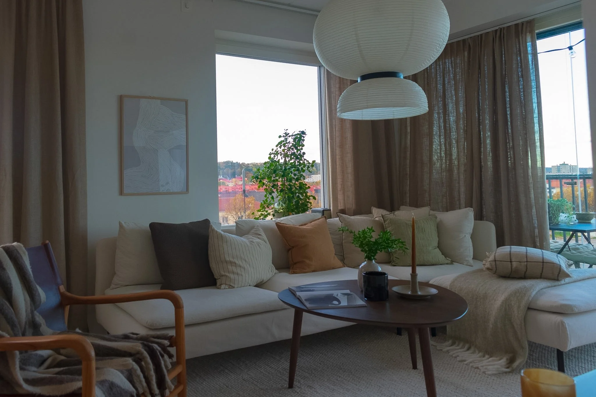 Living room with white sectional sofa, throw pillows, round coffee table, plants near large windows with beige curtains, pendant light overhead, city view outside.
