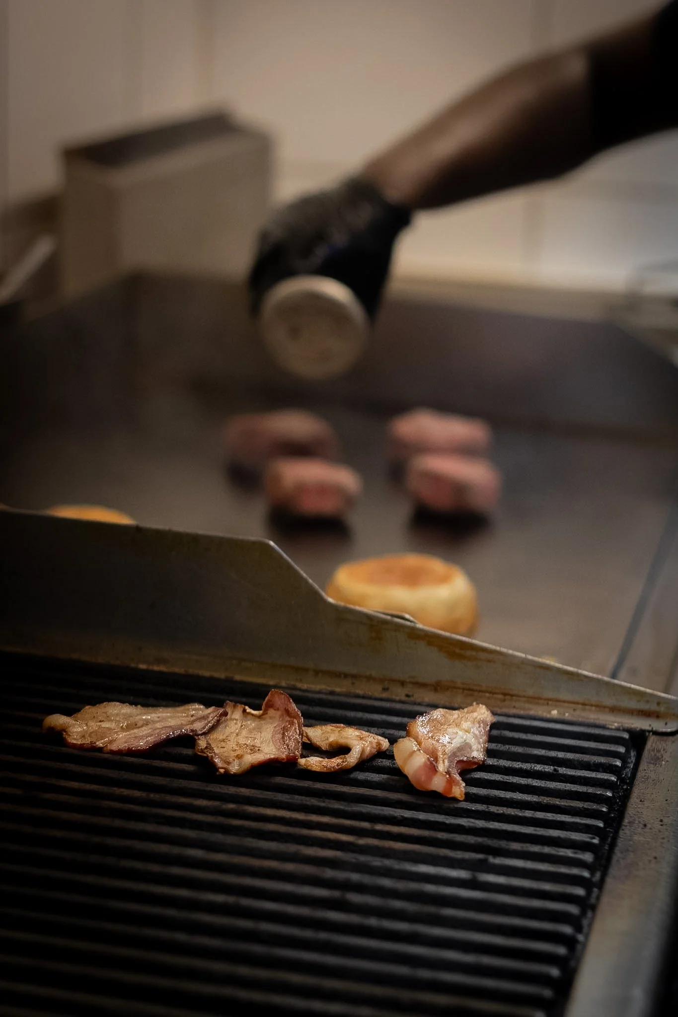 Cooking bacon and meat slices on a grill with a kitchen countertop and oven in the background.