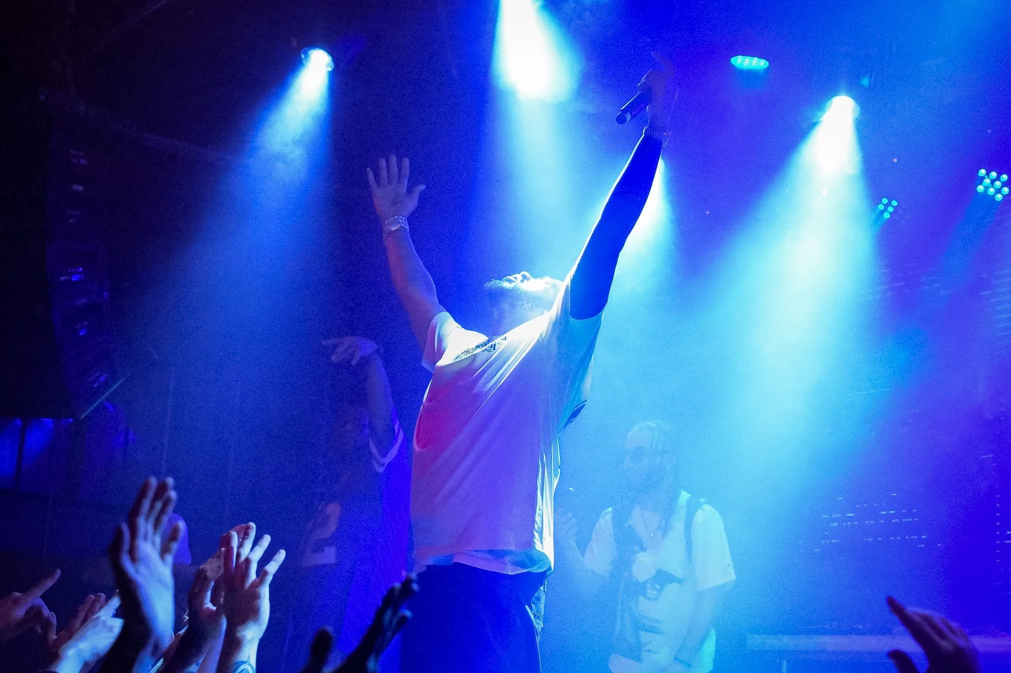 A performer on stage holding a microphone, with blue stage lighting and a crowd reaching out with hands.