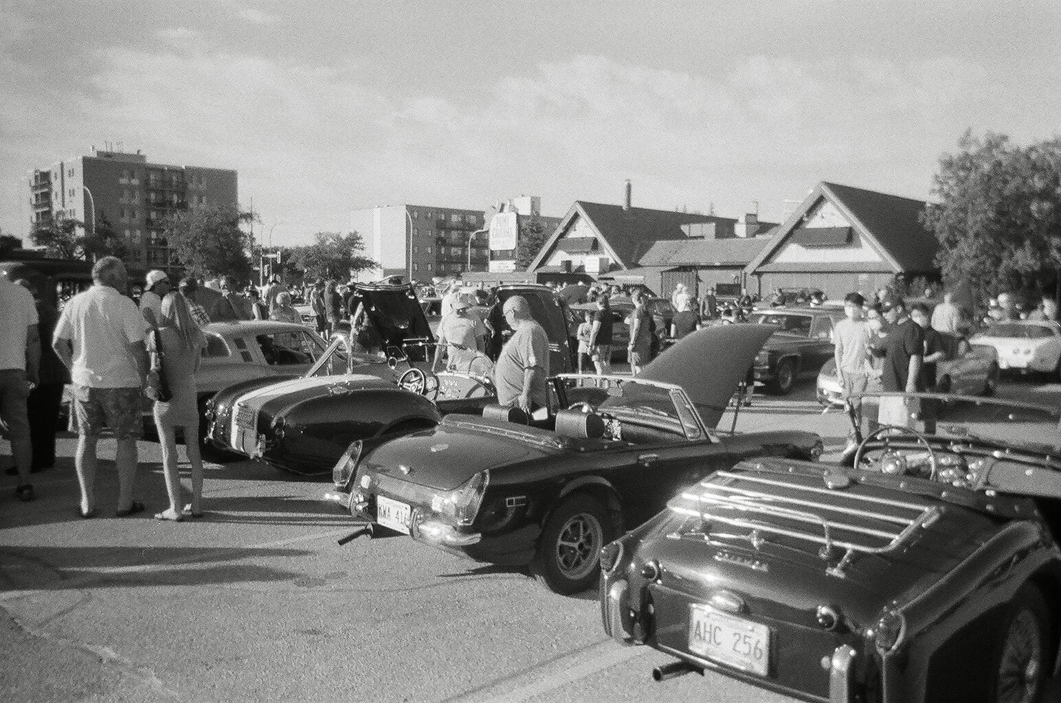 Black and white photo of a classic car show in a parking lot, with various vintage cars and a crowd of people admiring them.