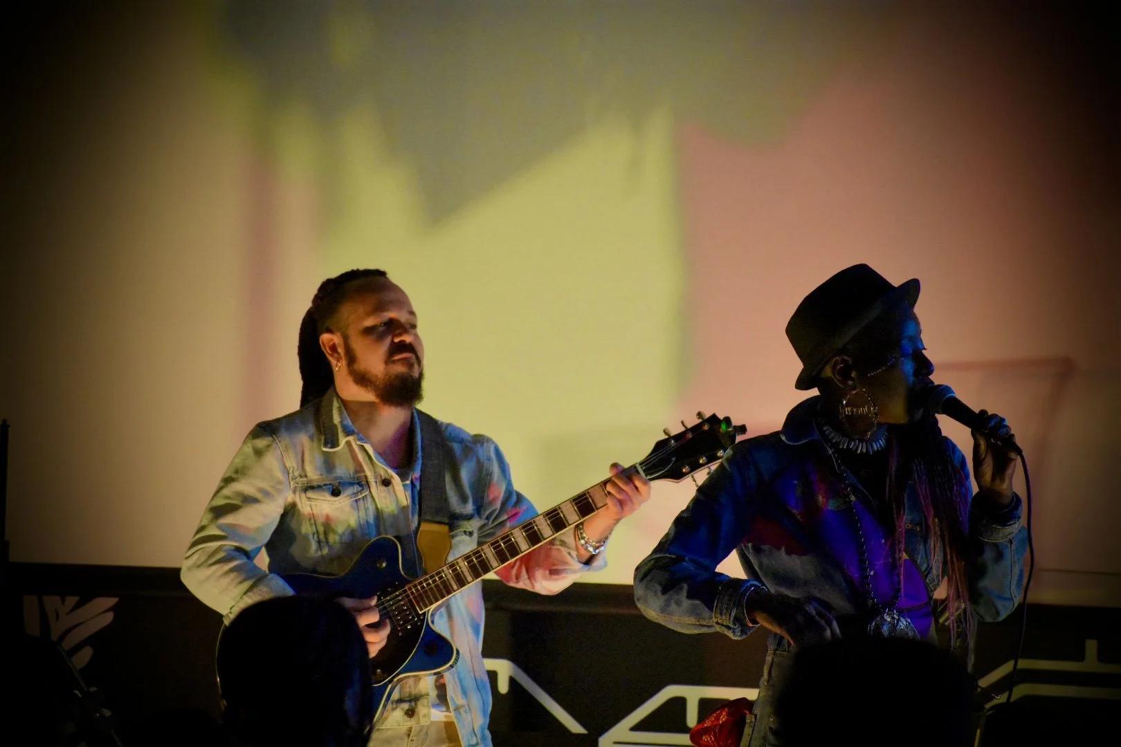 Two musicians on stage, one playing an acoustic guitar and the other singing into a microphone, with colorful lighting and a blurry background.