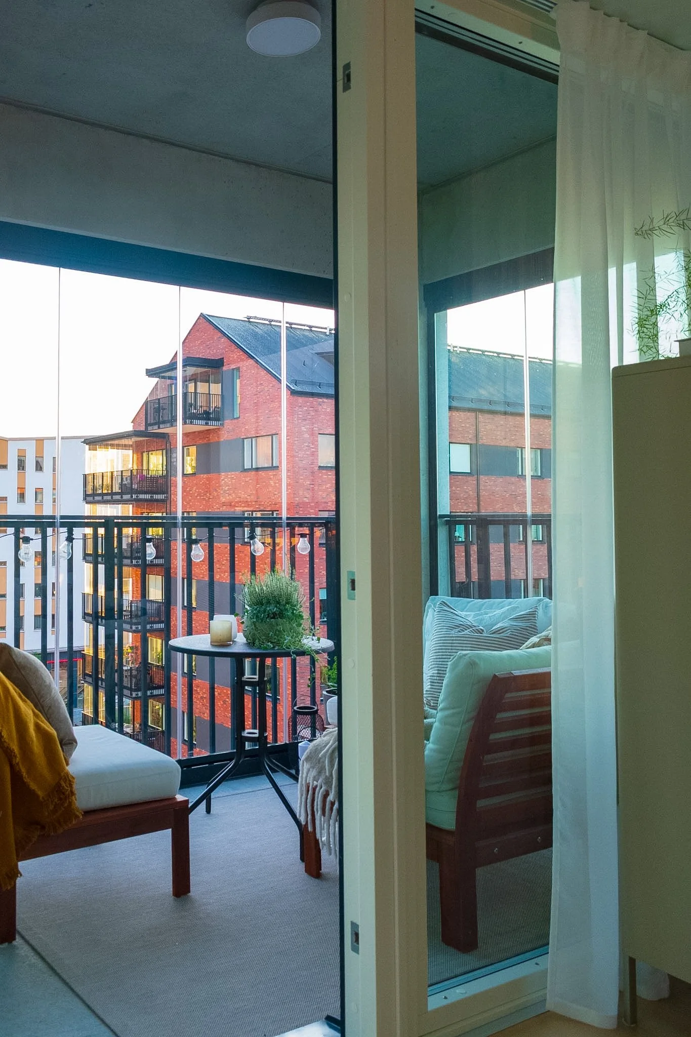 View of a balcony with outdoor seating, overlooking modern brick and white apartment buildings, with string lights hanging over the balcony railing, seen from inside a living room through sliding glass doors with sheer curtains and a white upholstere