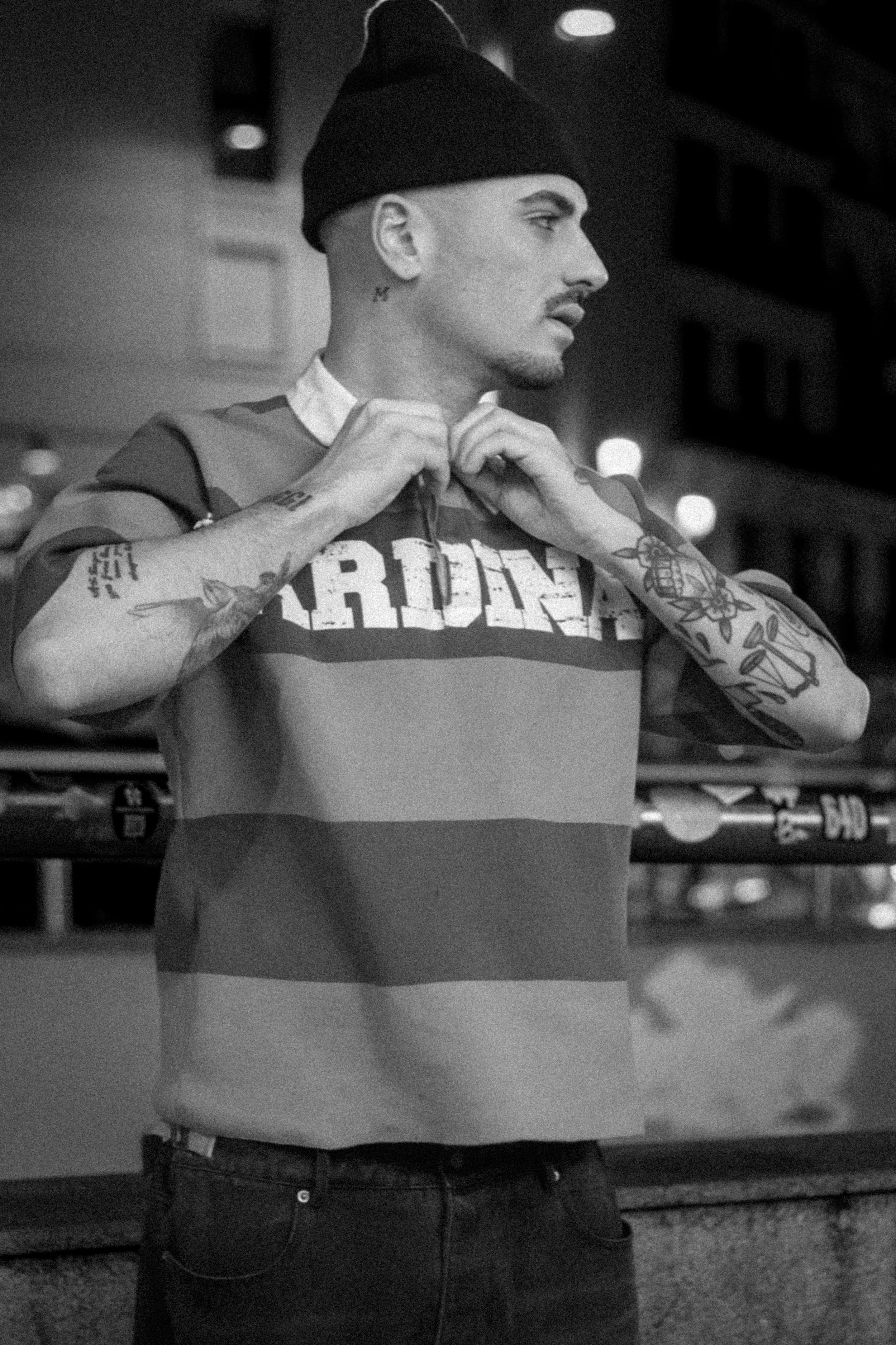A black and white photo of a man adjusting his hoodie at a bar or club, with tattoos on his arms and a tattoo near his neck, wearing a beanie and jeans.