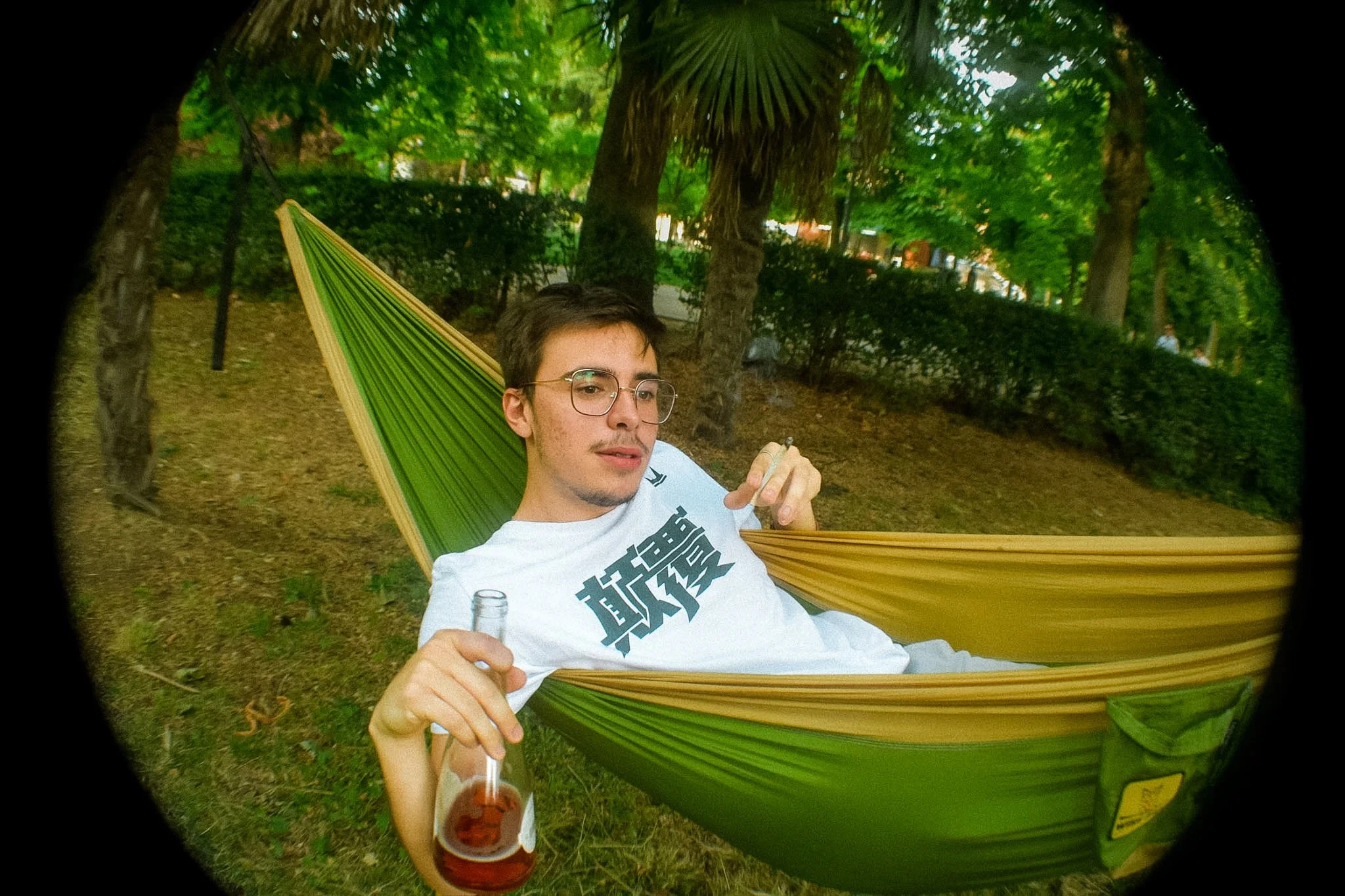 A young man with glasses relaxing in a hammock in a park, holding a glass of beer.