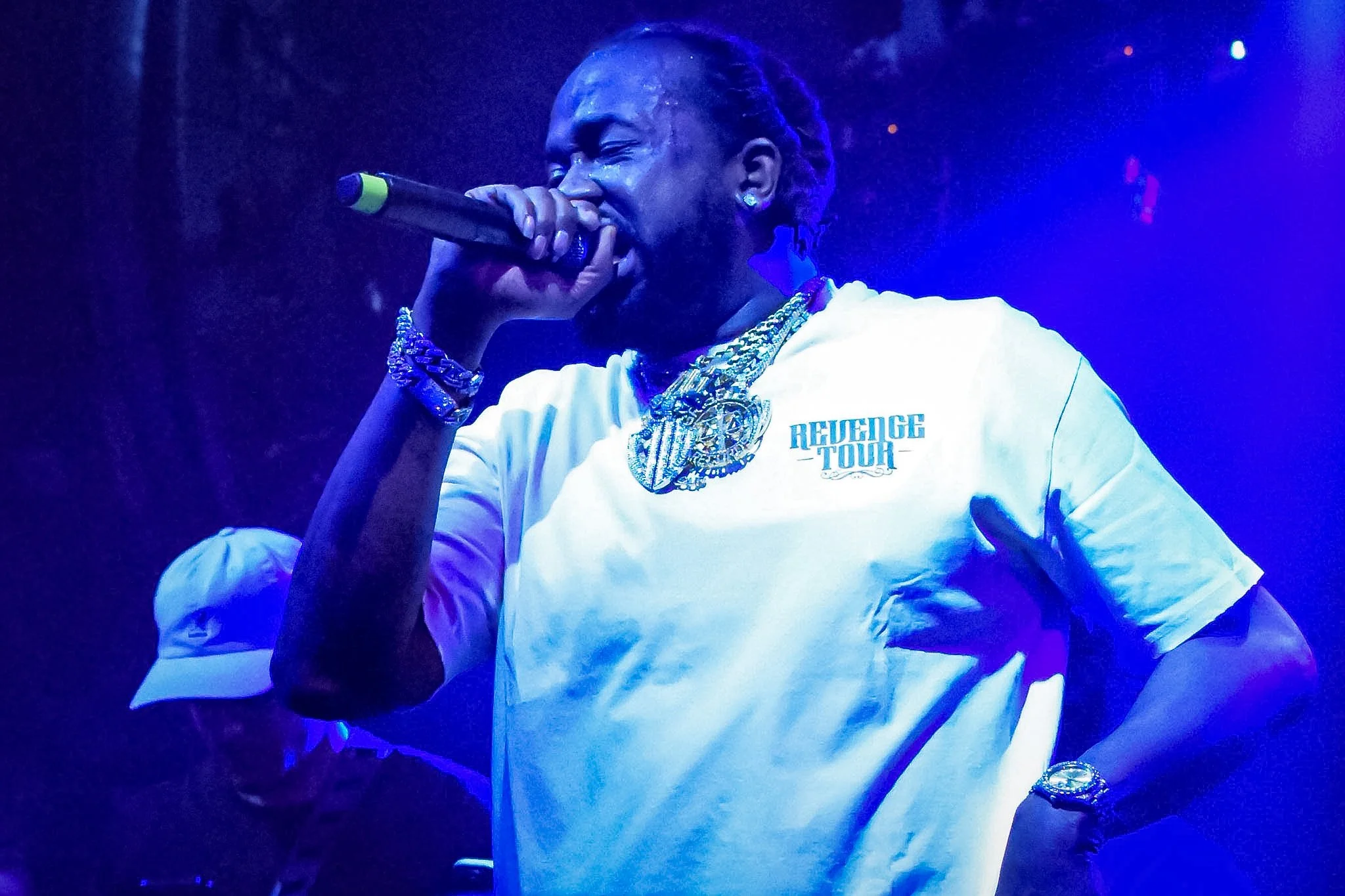 A person holding a microphone on stage, wearing a white shirt with 'Revenge Tour' printed on it, adorned with heavy jewelry, in a blue-lit environment.