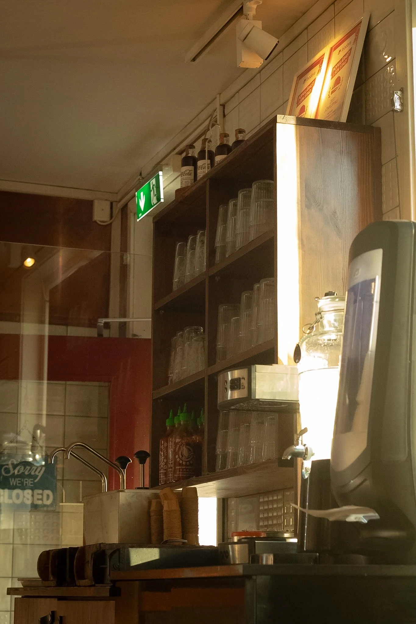 Interior of a small restaurant or café with wooden shelves holding glasses, bottles, and condiments. There is a microwave on the counter, and part of the kitchen setup with a sink and a 