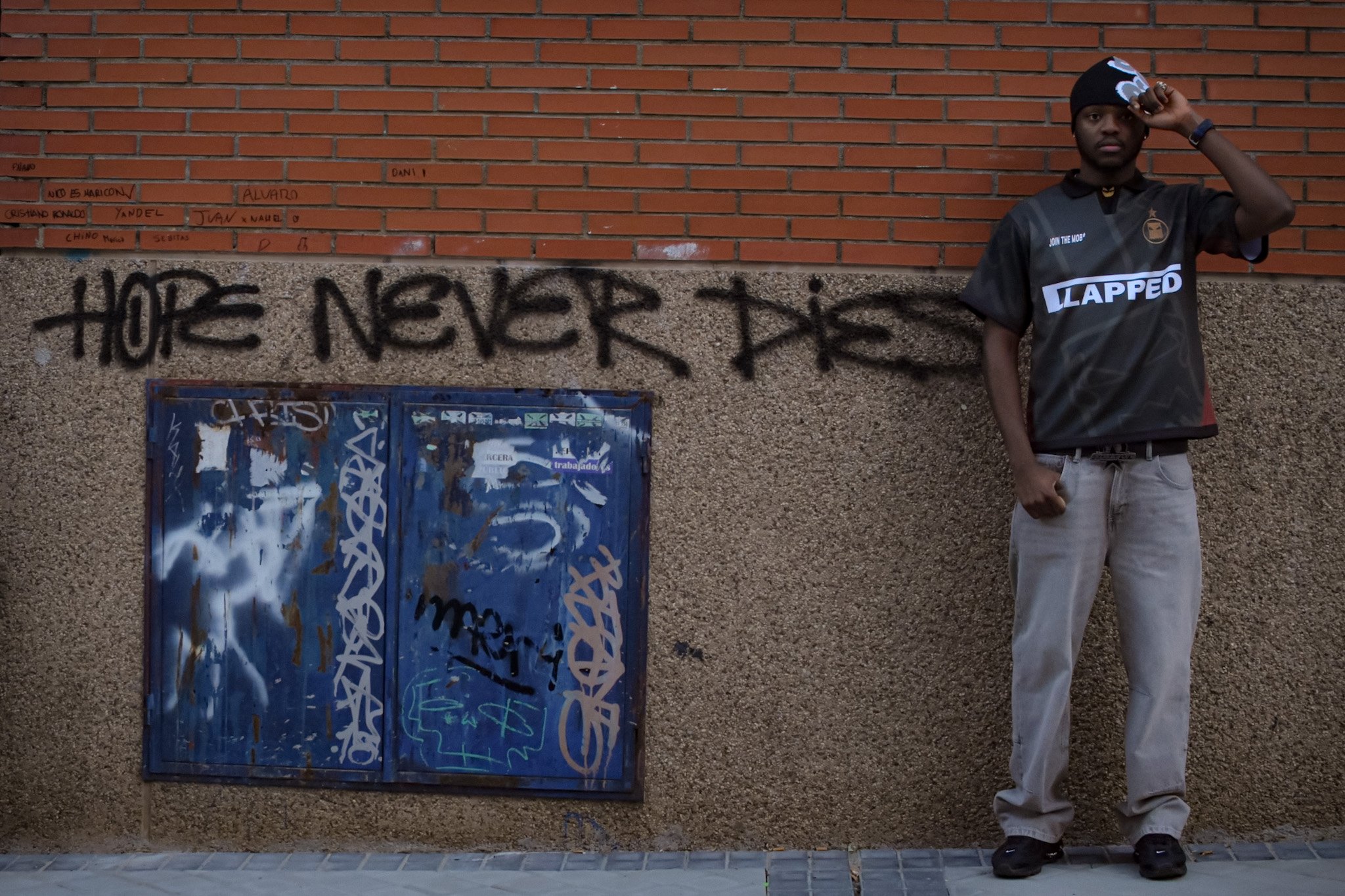 A young man standing next to a graffiti-covered wall with the words 'Hope Never Dies' spray-painted on it. He is wearing a black cap, a black and gray sports jersey with the phrase 'Join the Mob' and 'Lapped' on it, and light-colored jeans. He is tou