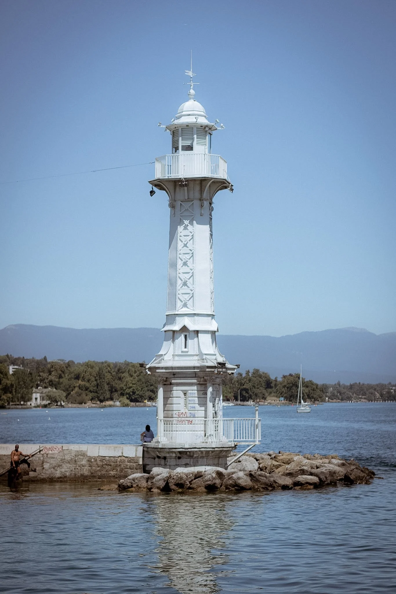 A white lighthouse situated by a body of water, with a sailboat and people nearby, mountains in the distance, and clear blue sky.