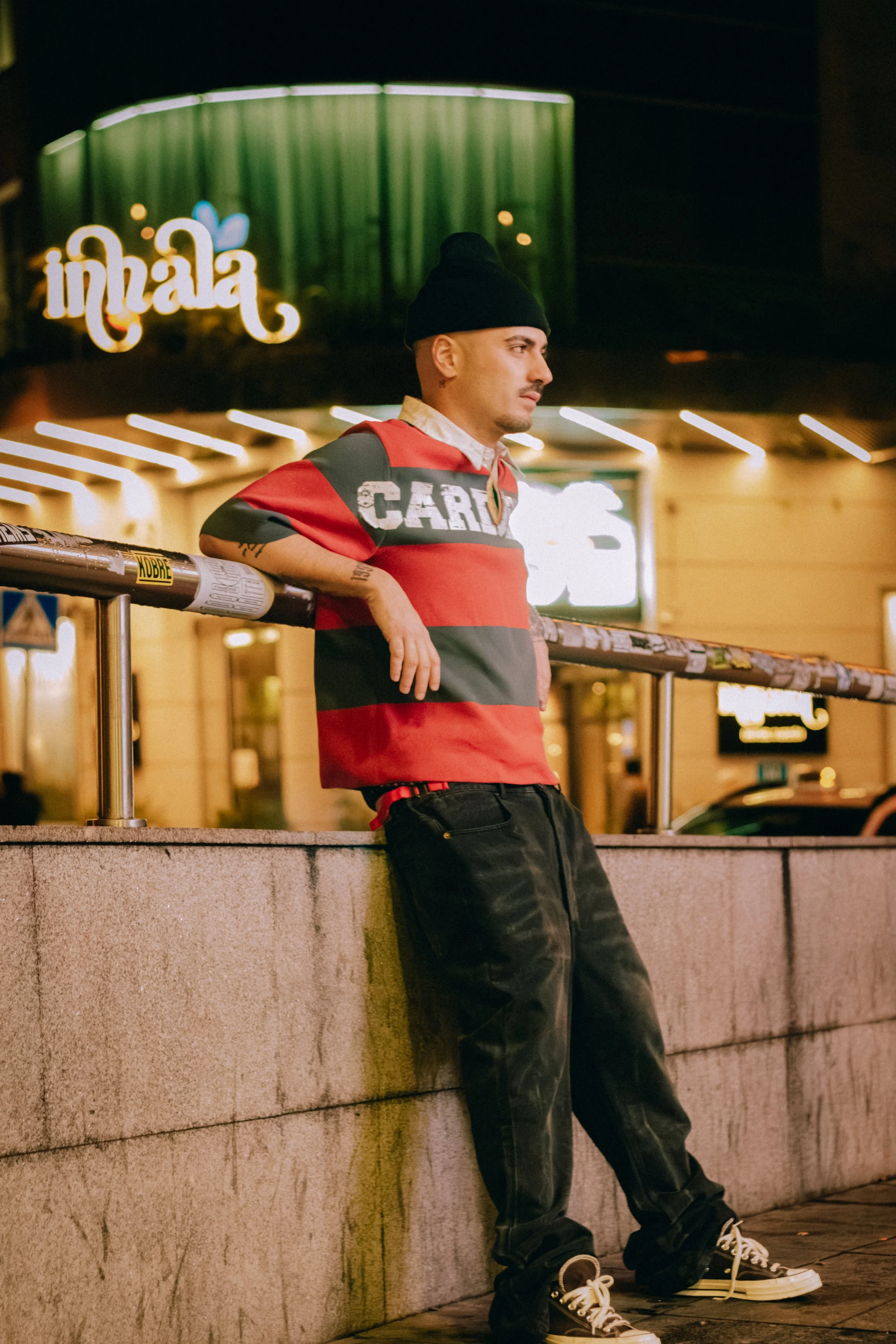 A man wearing a black beanie, a red and gray striped shirt, black pants, and sneakers leaning against a concrete wall at night, with illuminated signs and lights in the background.