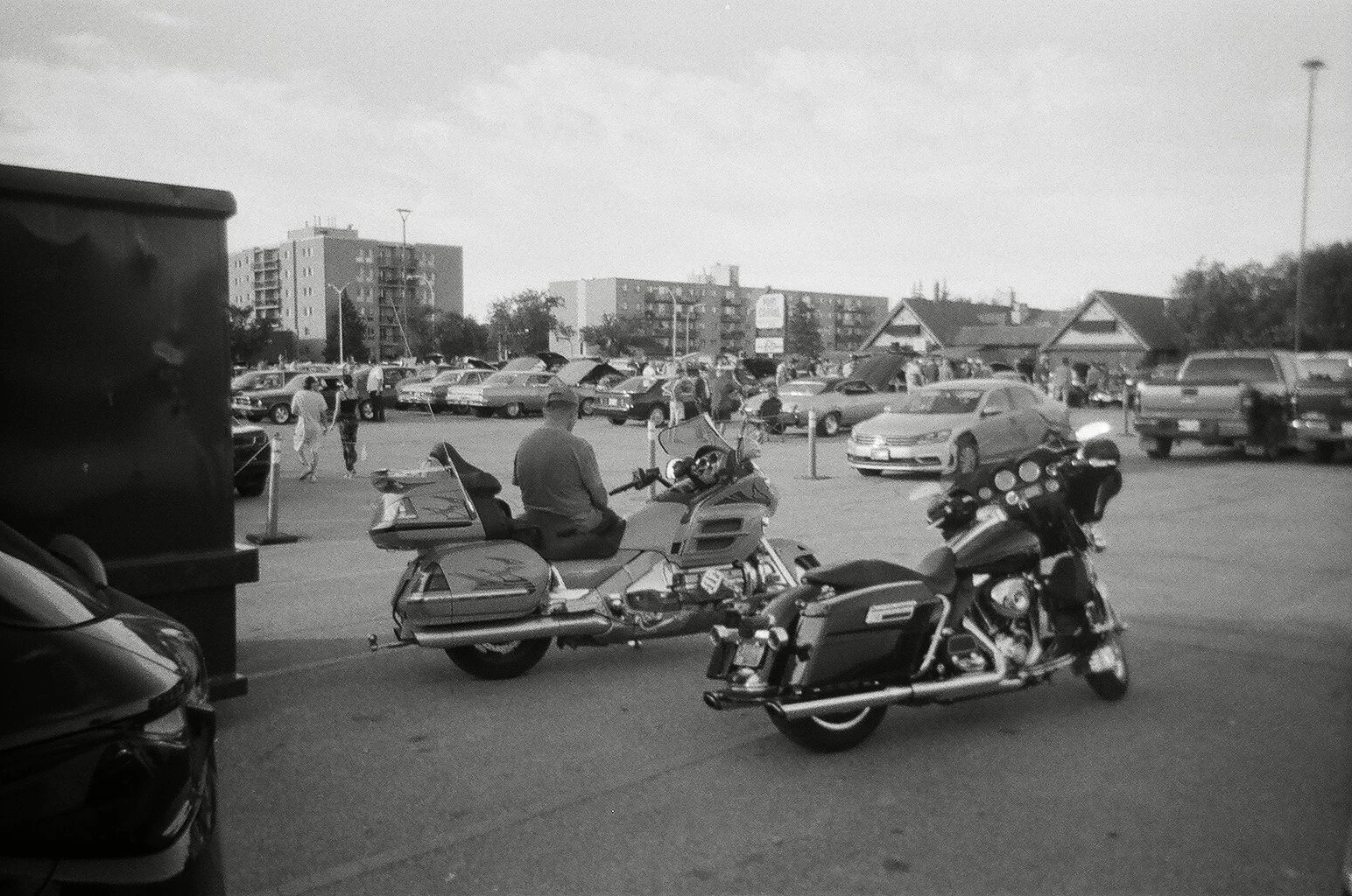 A parking lot with several parked cars and two motorcycles in the foreground, a man riding a motorcycle, and people walking around. In the background, there are multi-story apartments and small houses.
