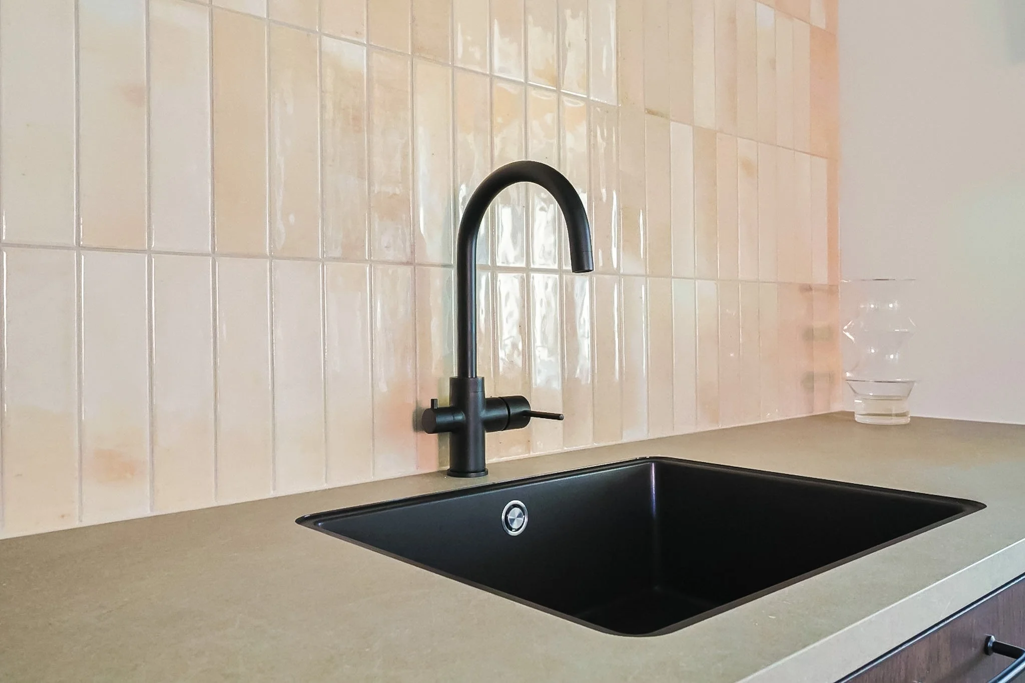A kitchen sink with a black faucet, beige tile backsplash, beige countertop, and a clear glass of water on the right side of the sink.