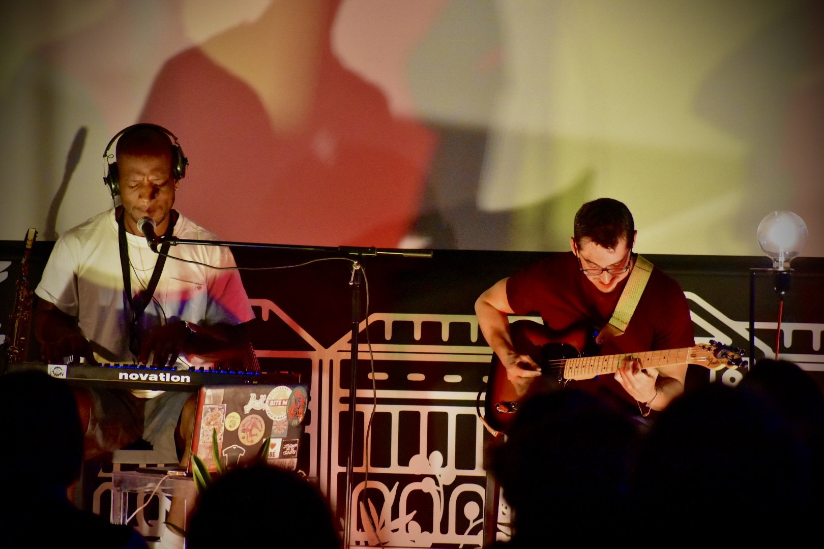 Two musicians performing on stage, one playing a keyboard and singing with headphones and a microphone, the other playing an electric guitar and smiling, with a large colorful abstract background projection.