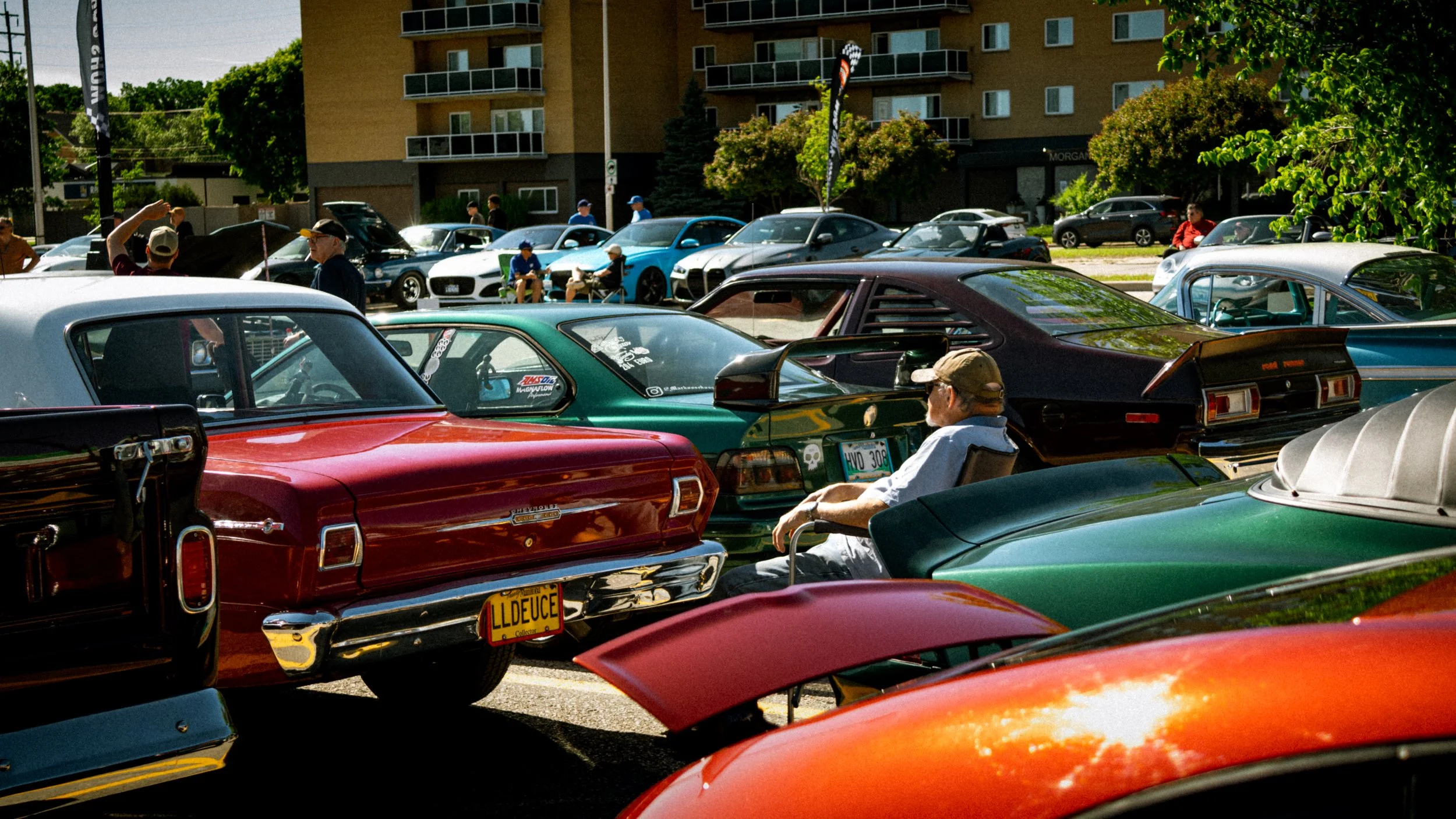 A crowded parking lot filled with vintage and classic cars, with people walking around and sitting in chairs, during a car show event on a sunny day. In the background, there is a multi-story apartment building and green trees.