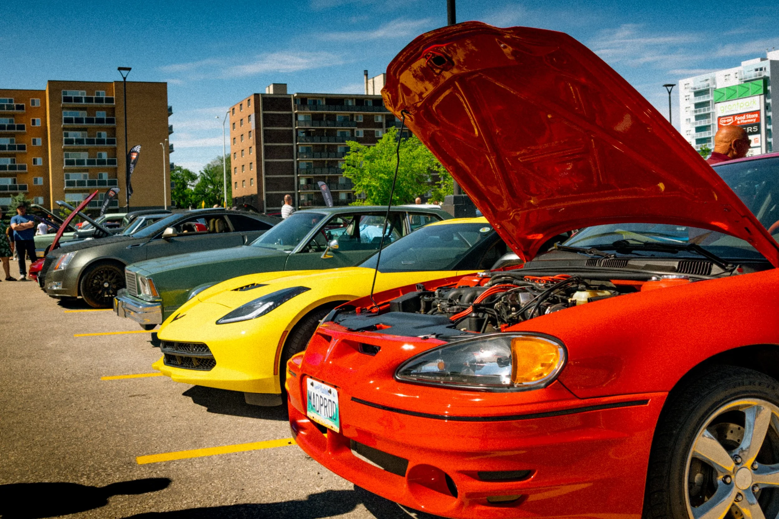 A row of sports cars parked in an outdoor lot during daytime, with their hoods open. There are buildings and a few people in the background.