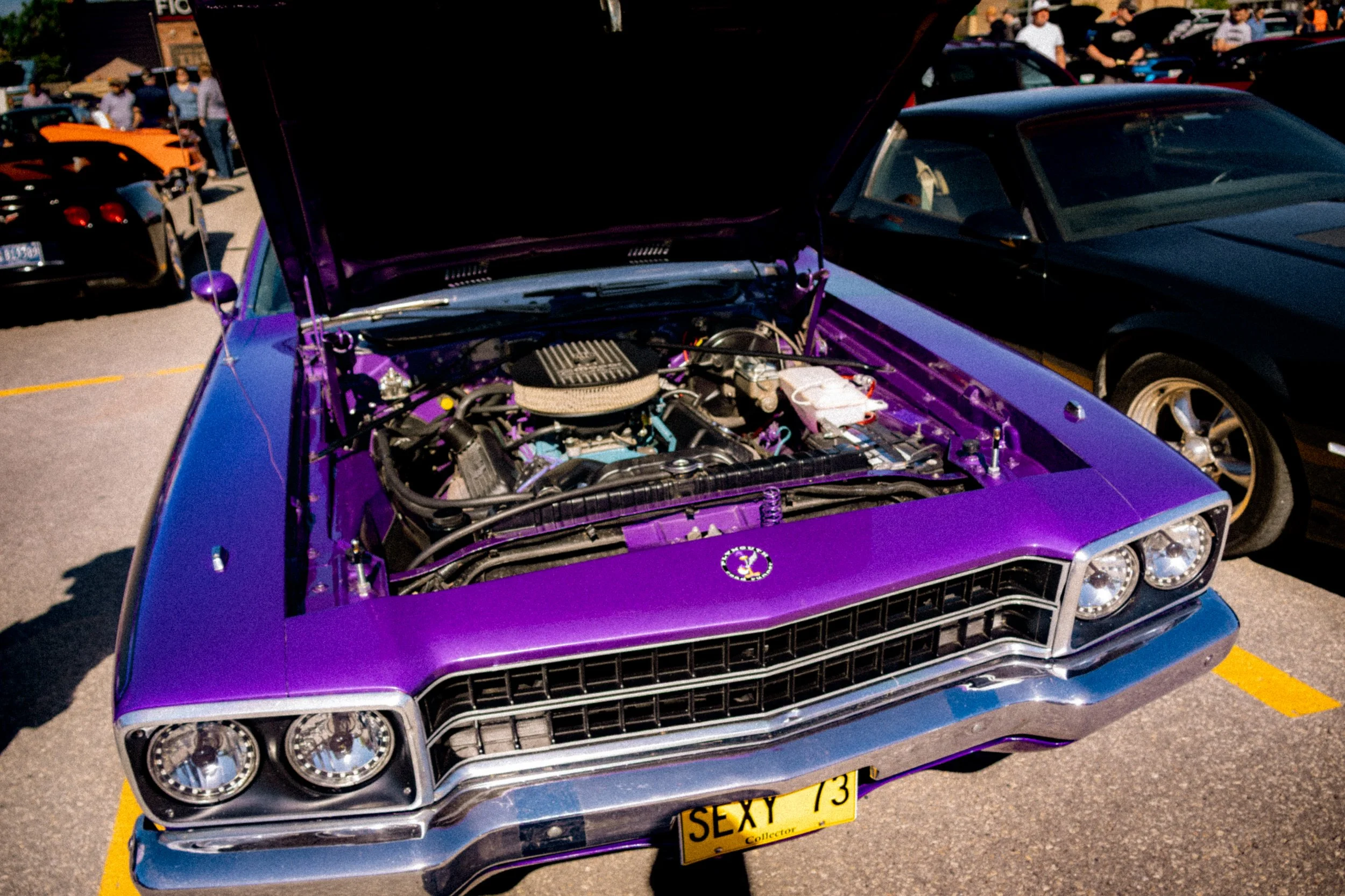 A purple classic car with its hood open, showing the engine, parked among other cars at a car show, with spectators in the background.