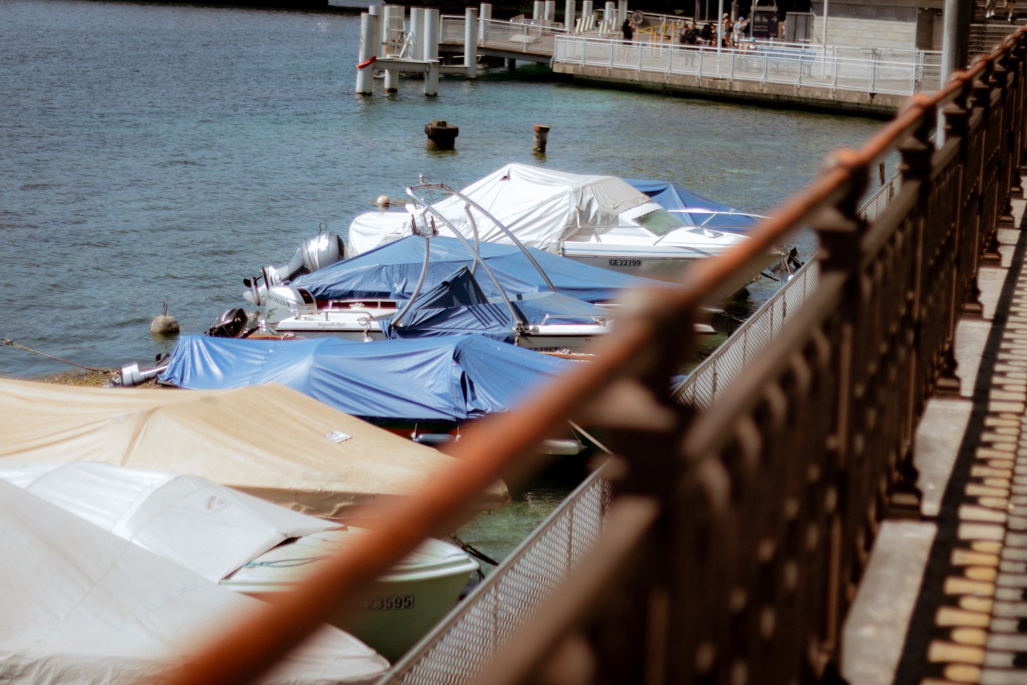 Boats docked along a pier with a railing, some boats covered with tarps, and people sitting on an upper deck in the background.