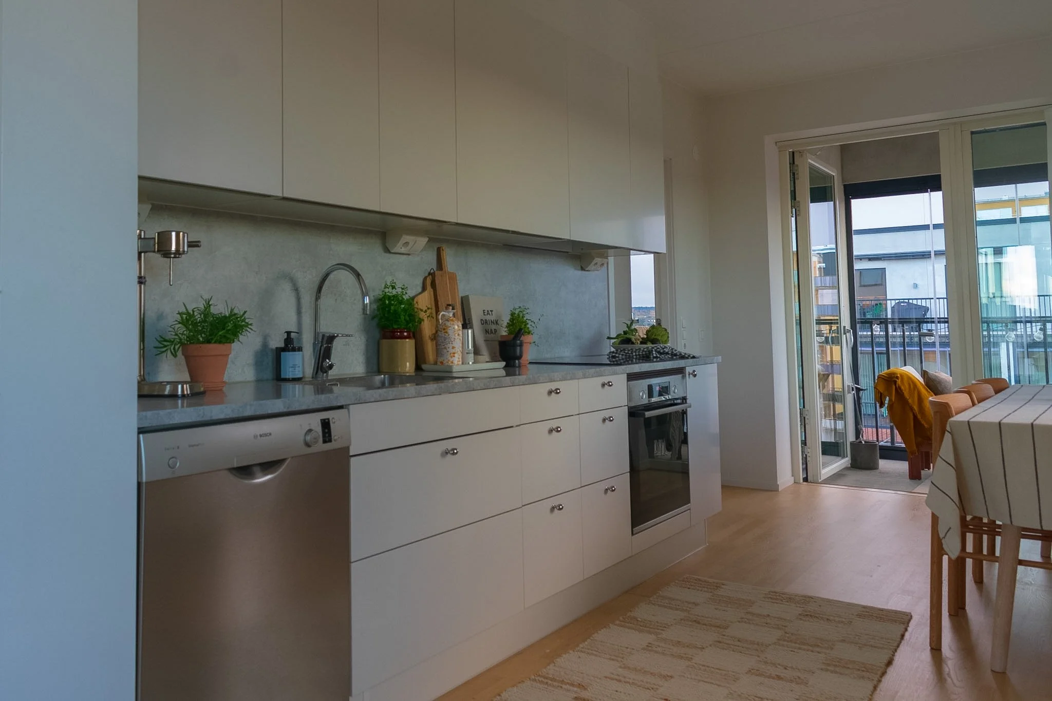 Modern kitchen with white cabinets, a marble countertop, and a backsplash. There are potted herbs, a soap dispenser, a cutting board, and a small plant on the counter. A dishwasher and an oven are built into the cabinets. To the right, a dining table