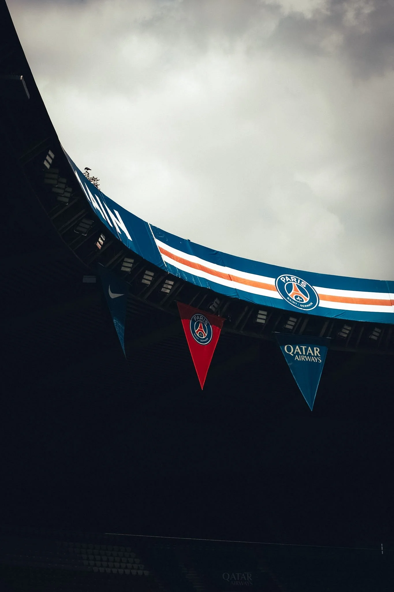 View of a stadium with banners featuring the Paris Saint-Germain football club logo, Qatar Airways sponsorship, and the Nike logo, with overcast sky above.