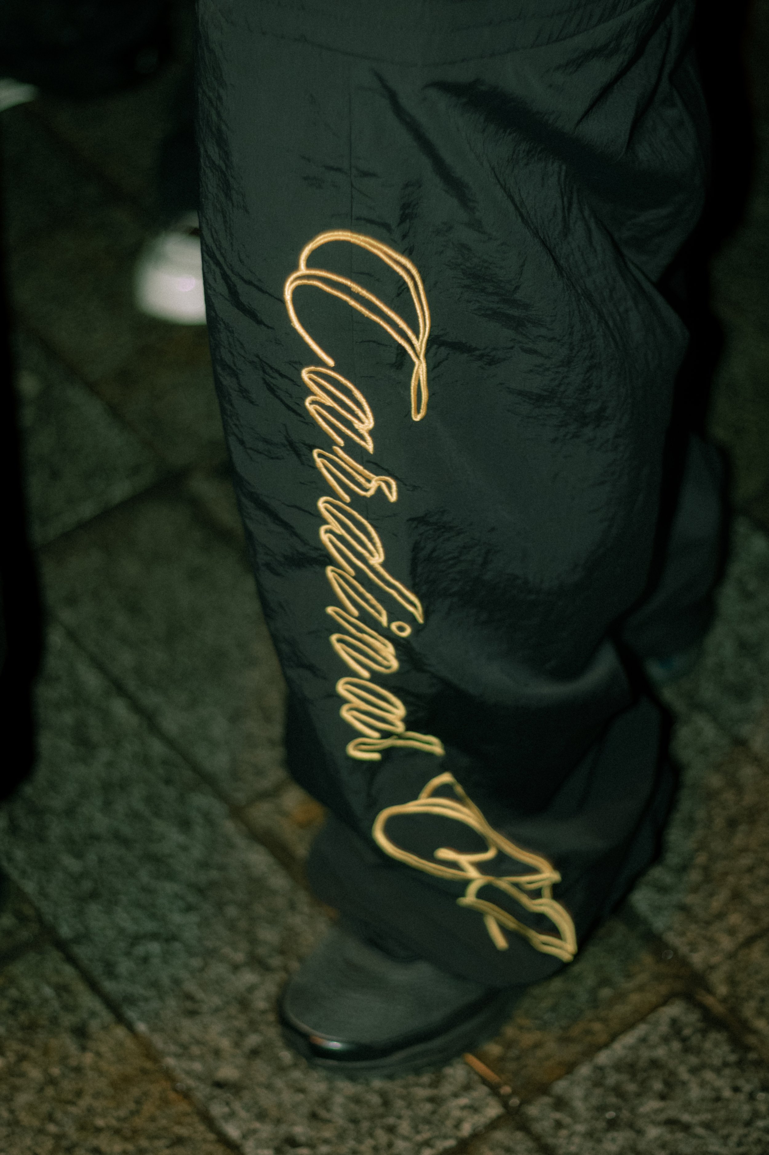 Close-up of a person's lower body wearing black athletic pants with a yellow script logo down the side, standing on a tiled floor.