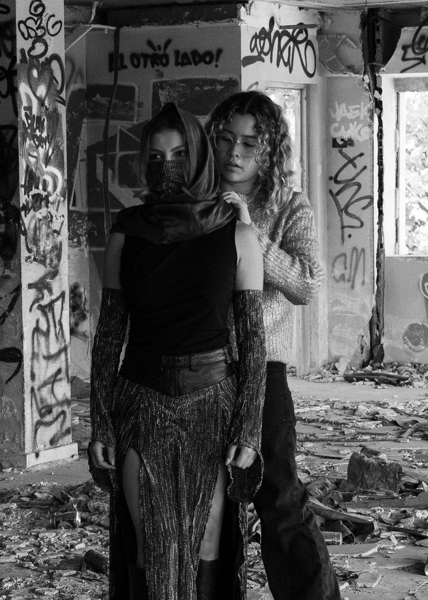 Two women stand close together in a dilapidated, graffiti-covered building; one woman wears a mask and a hood, the other has curly hair and glasses, both dressed in fashionable, shiny clothing.