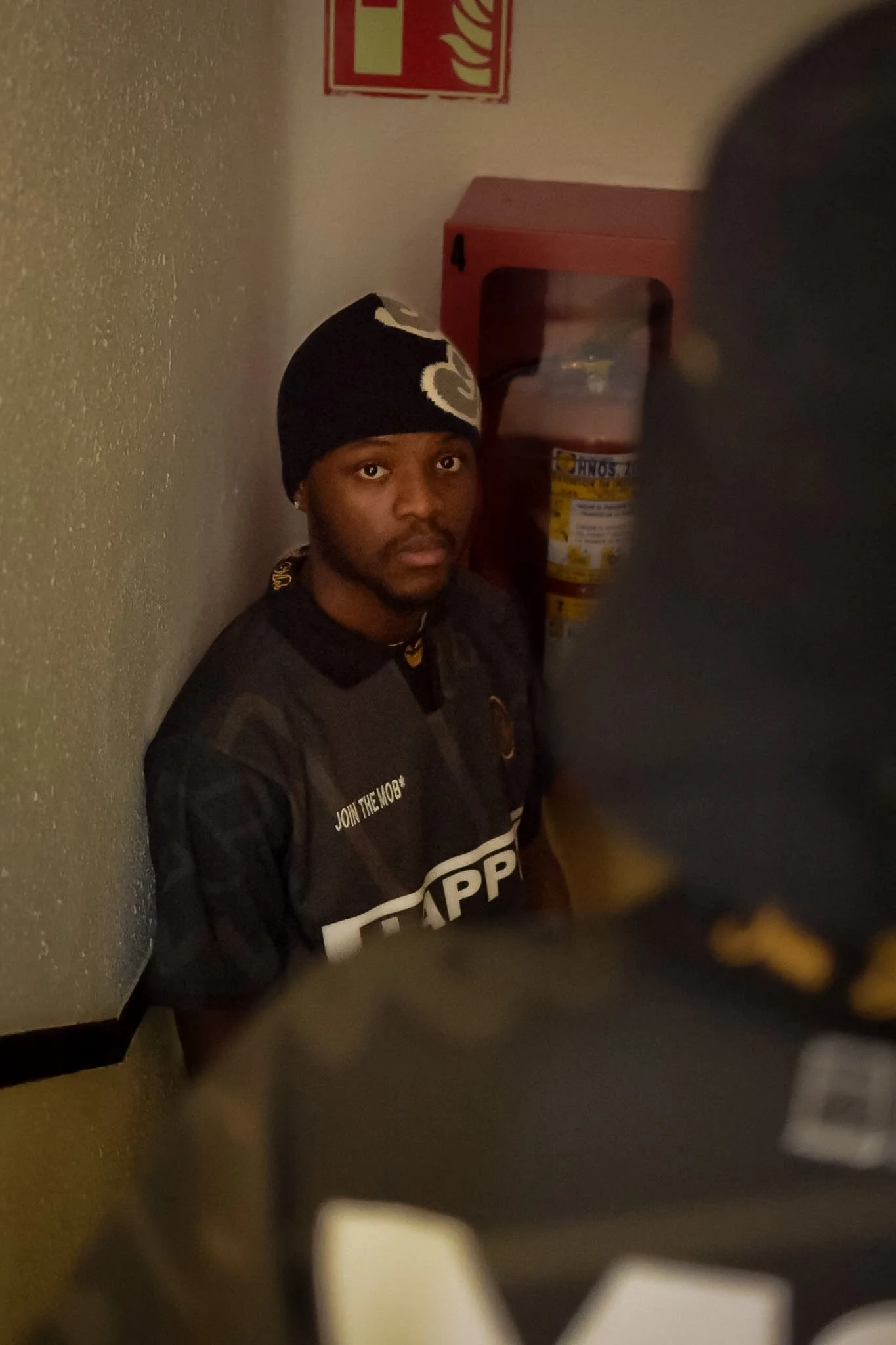 A young man with a black beanie and a black and white hoodie, standing in front of a beige textured wall, near a fire alarm and a red cabinet, indoors.