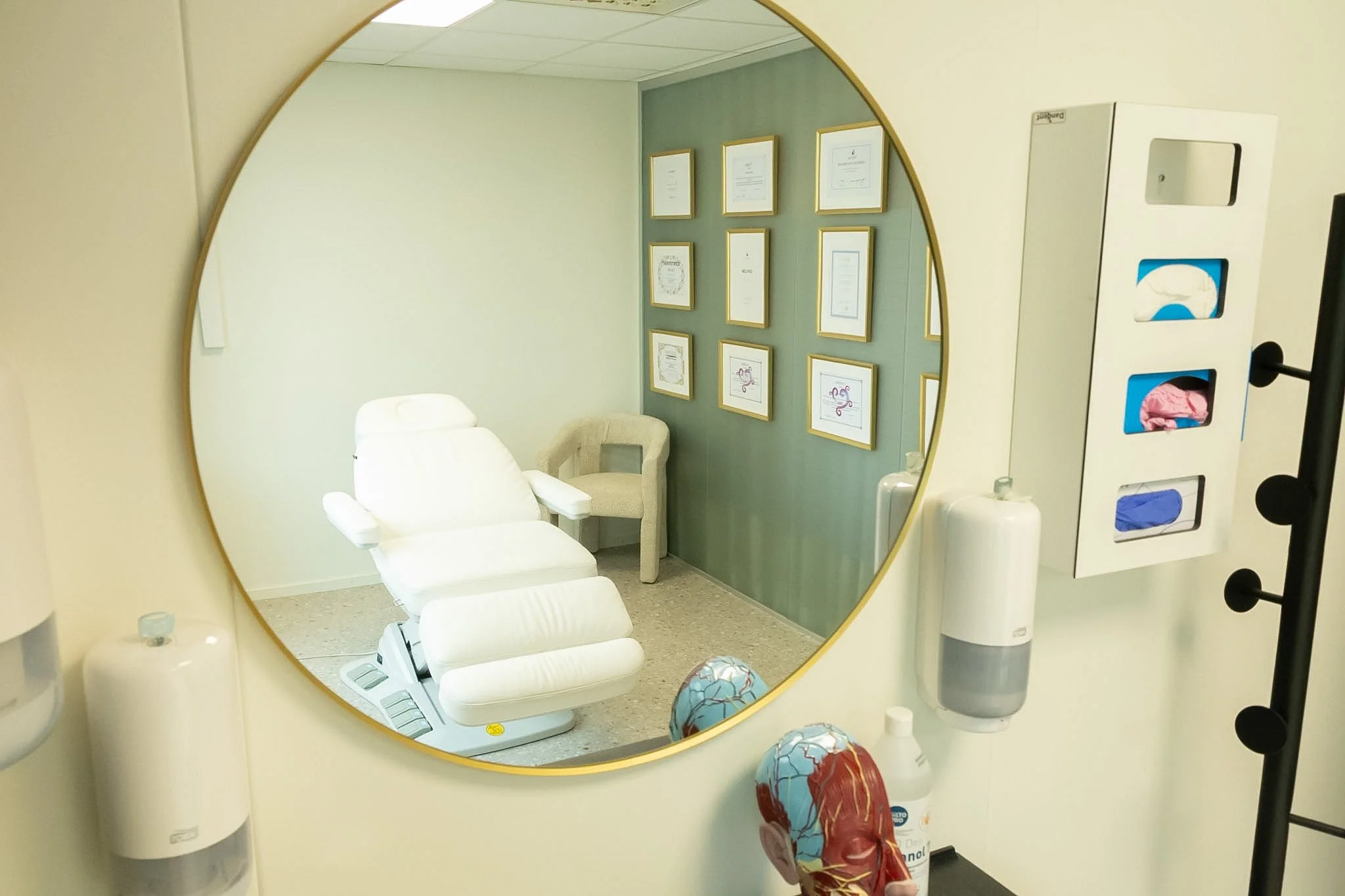 Hospital or medical office examination room seen through a round mirror. The room has an examination table, framed certificates or awards on the wall, and medical supplies like gloves, hand sanitizer, and a tissue box.