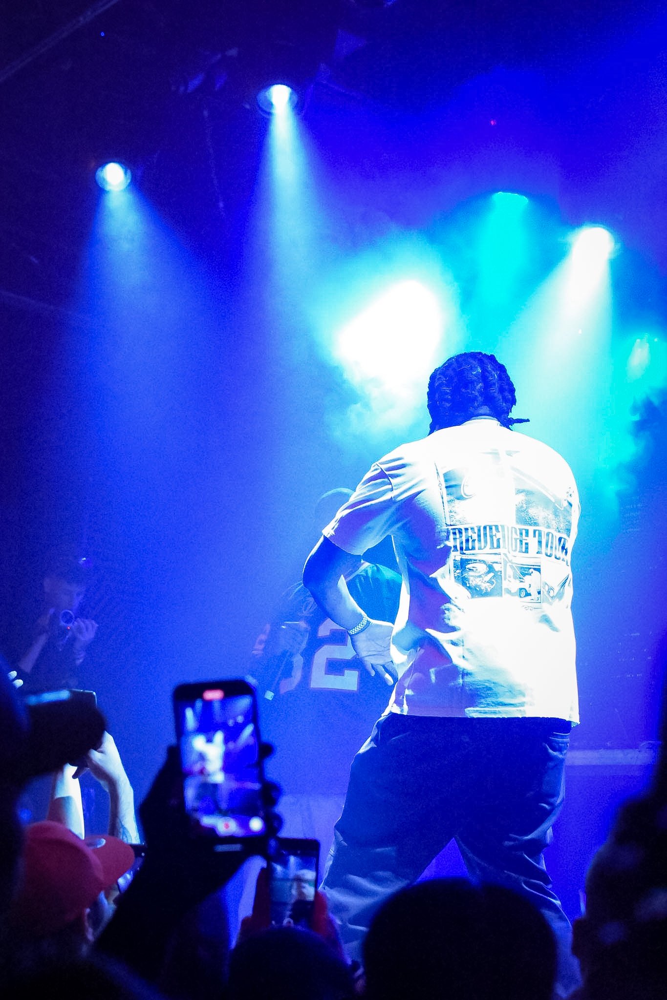 A person in a white jacket performing on stage under blue lights, with an audience taking photos in front.
