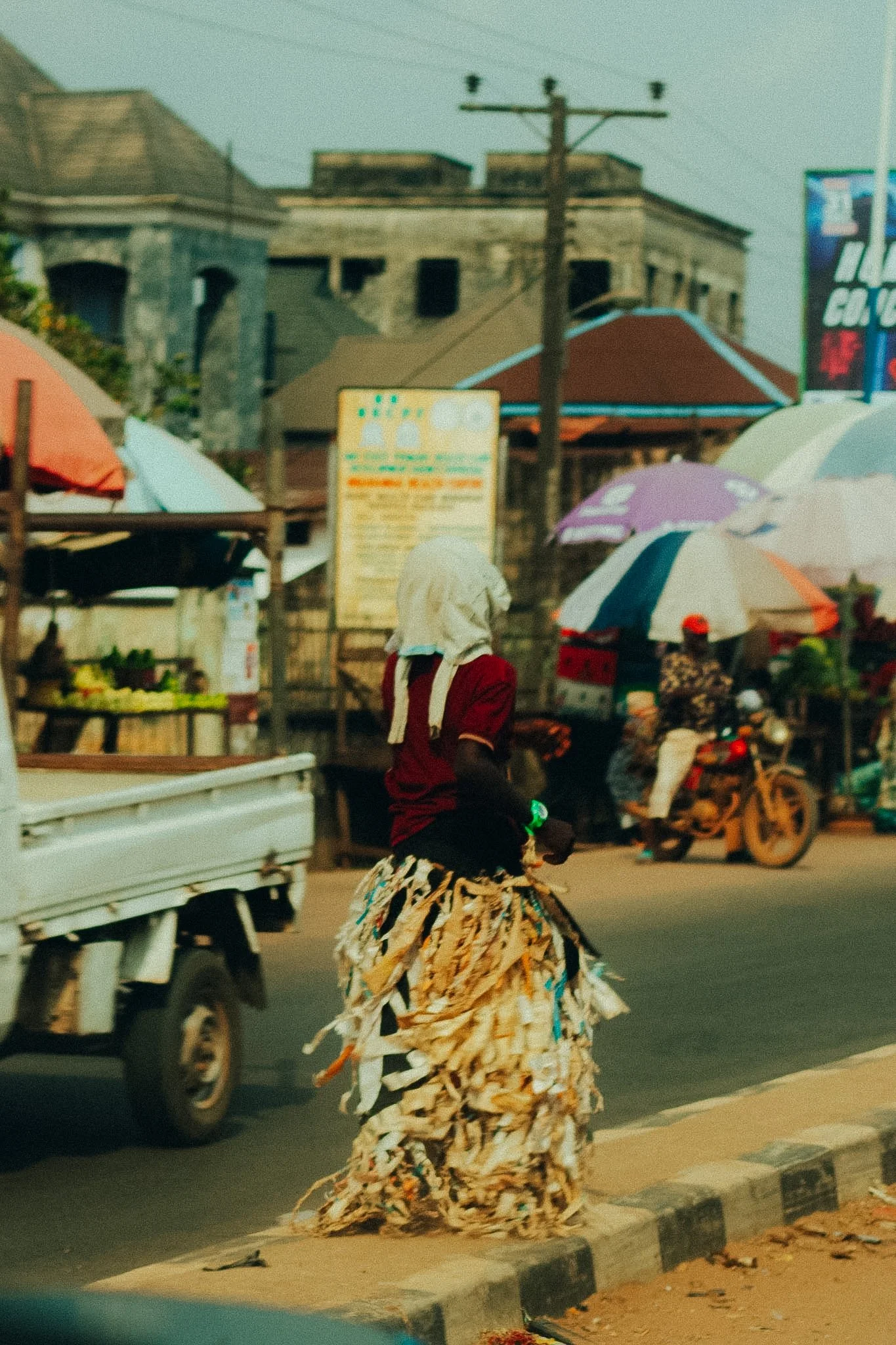 A person in traditional attire with a long skirt made of torn fabric walking on the street in an urban area with buildings, walls, and parked vehicles, including umbrellas providing shade to street vendors.