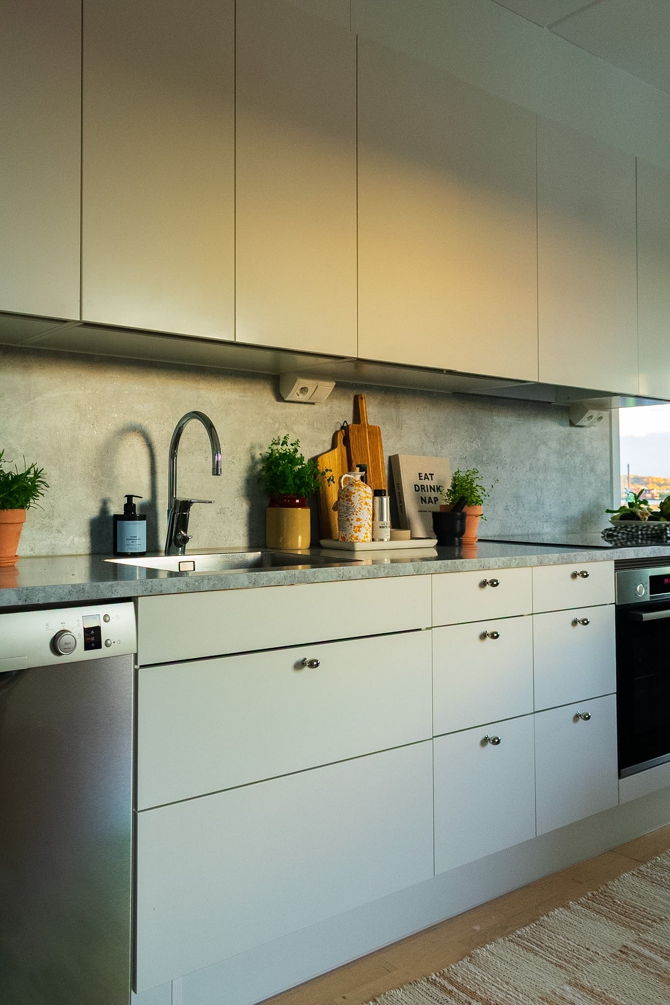 Modern kitchen with white cabinets, a sink with a faucet, plants, a wooden cutting board, and kitchen essentials on the countertop. There is a window showing an outdoor view.