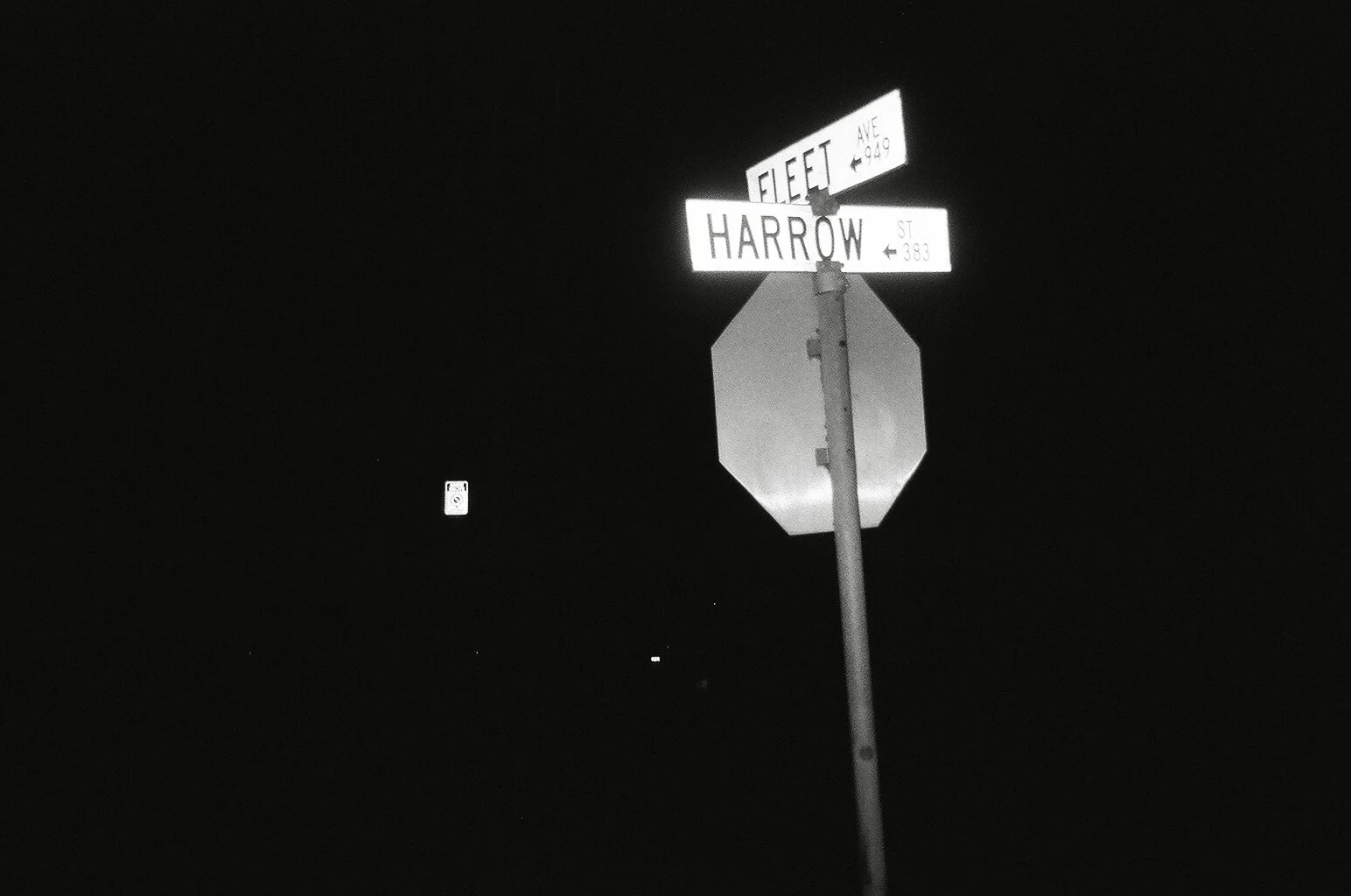 Street signs at night indicating the intersection of Fleet Avenue and Harrow Street, with a dark sky and minimal visible background.