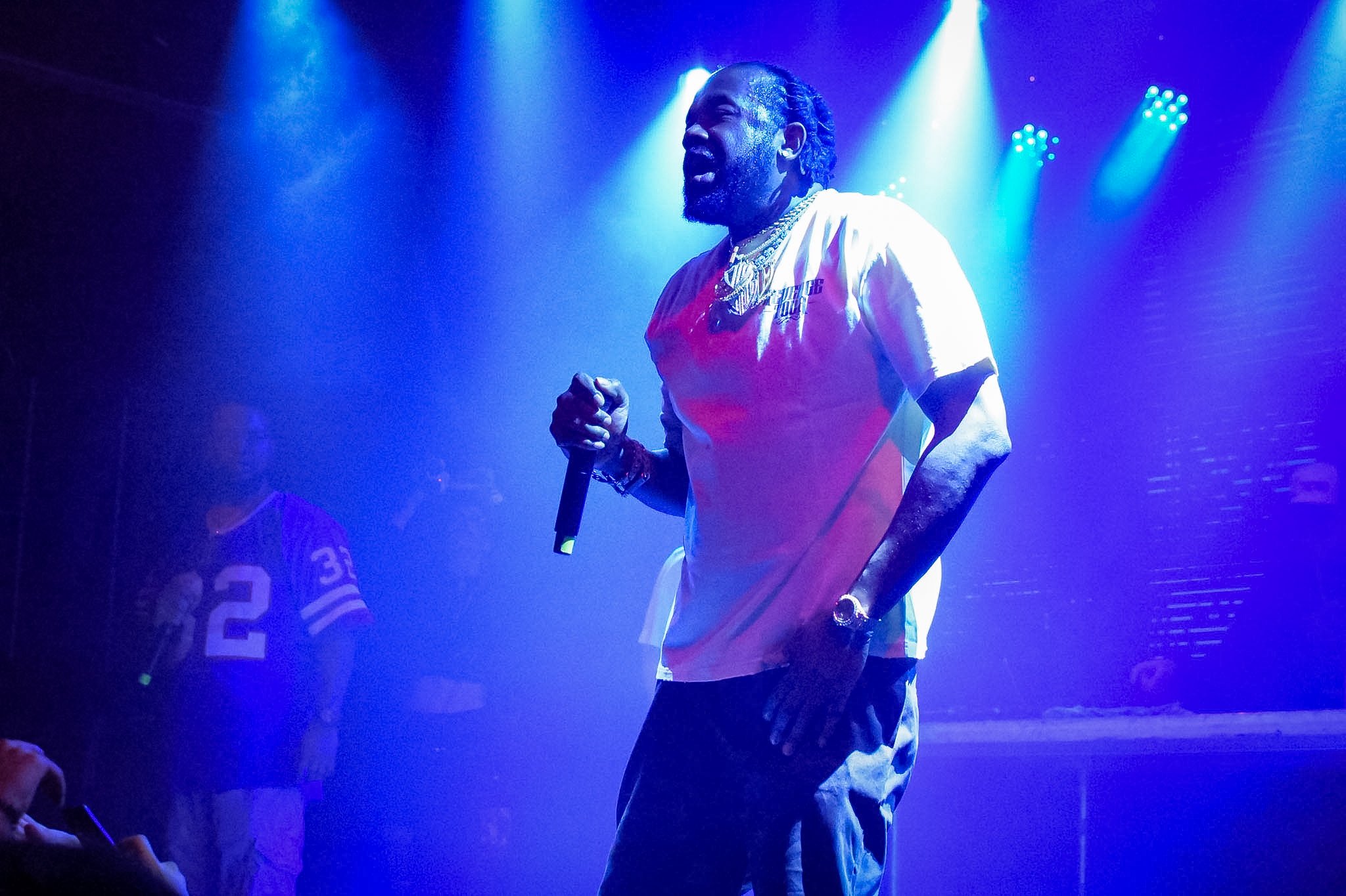 A man is performing on stage with blue lighting, holding a microphone, wearing a white T-shirt with red sleeves, and accessorized with necklaces and a gold watch.