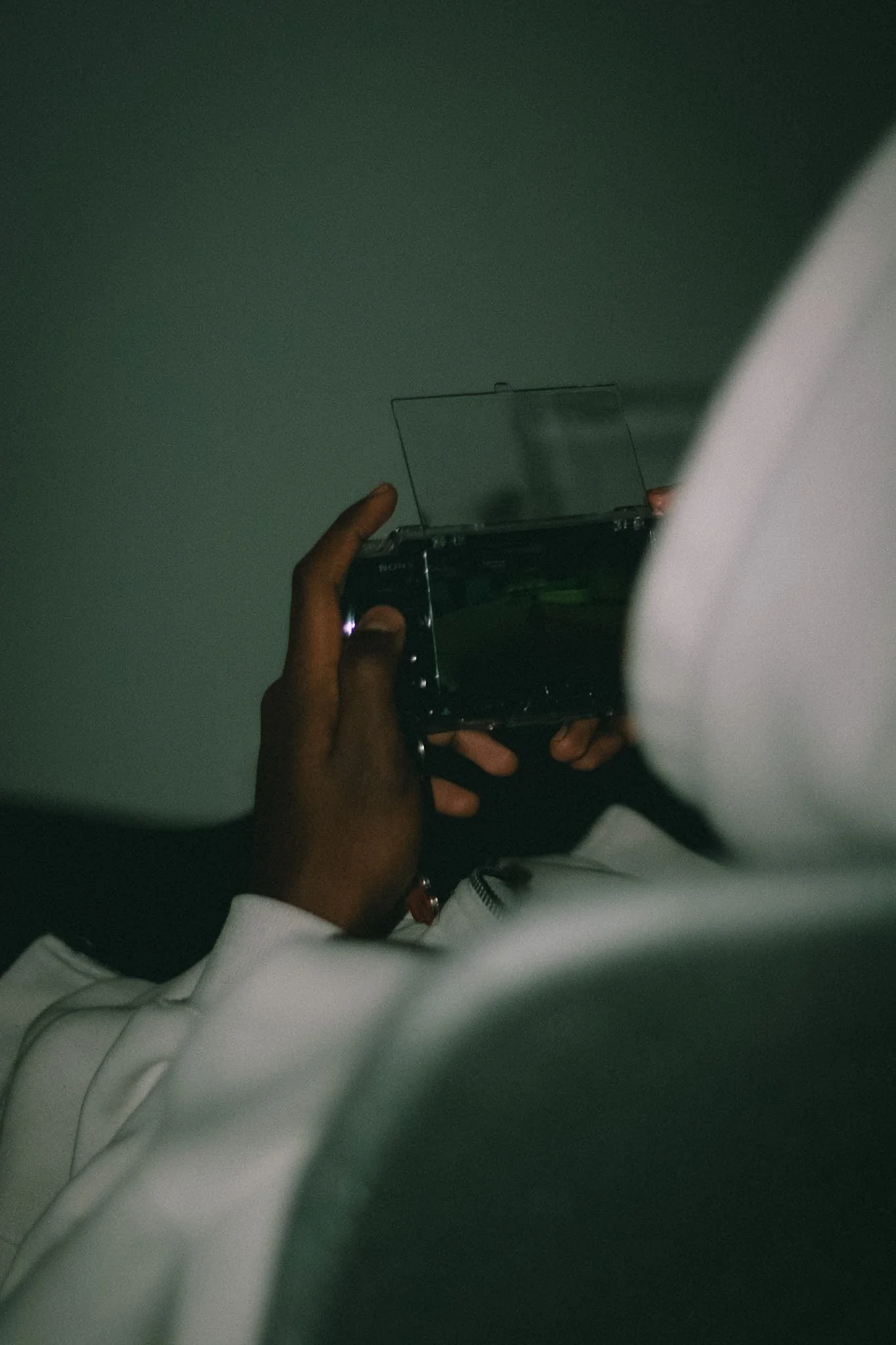 Person in white clothing taking a photo with a camera, with the focus on their hand and camera, and part of their face visible, against a greenish background.