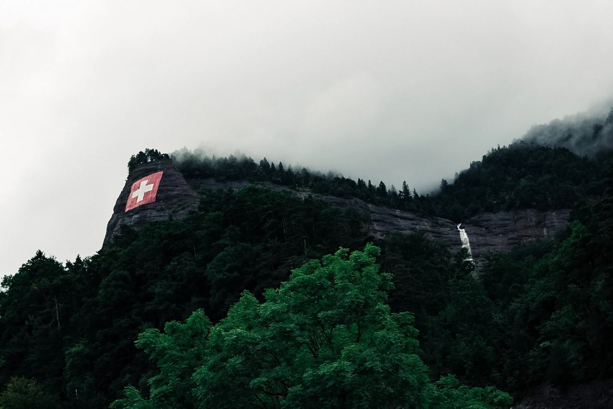 A mountain with a Swiss flag painted on a large rock formation with a waterfall cascading down nearby, surrounded by lush green trees and foggy weather.