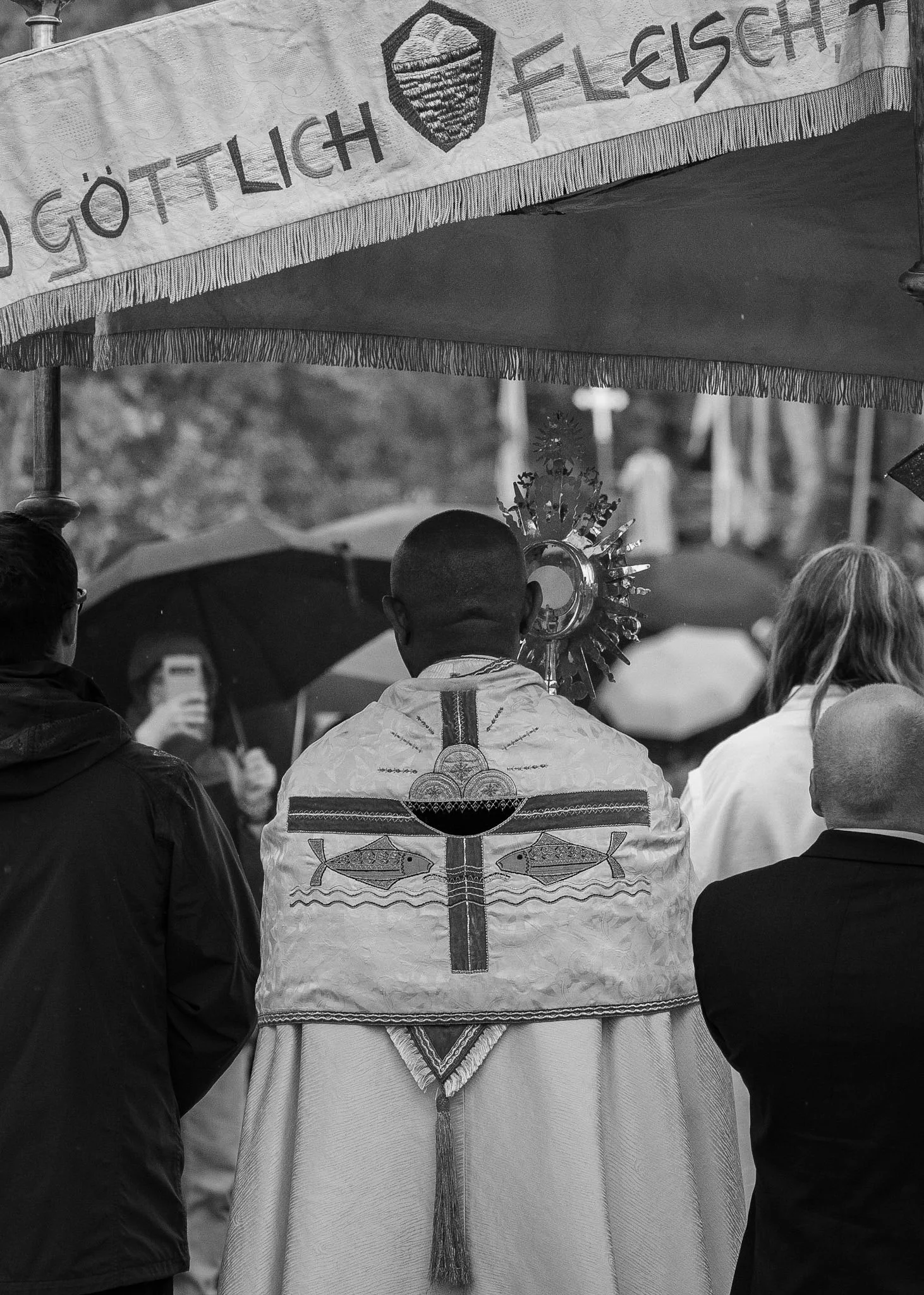 People standing together at a gathering, with one person wearing a traditional embroidered garment and holding a ceremonial object, in front of a banner with text in a European language.