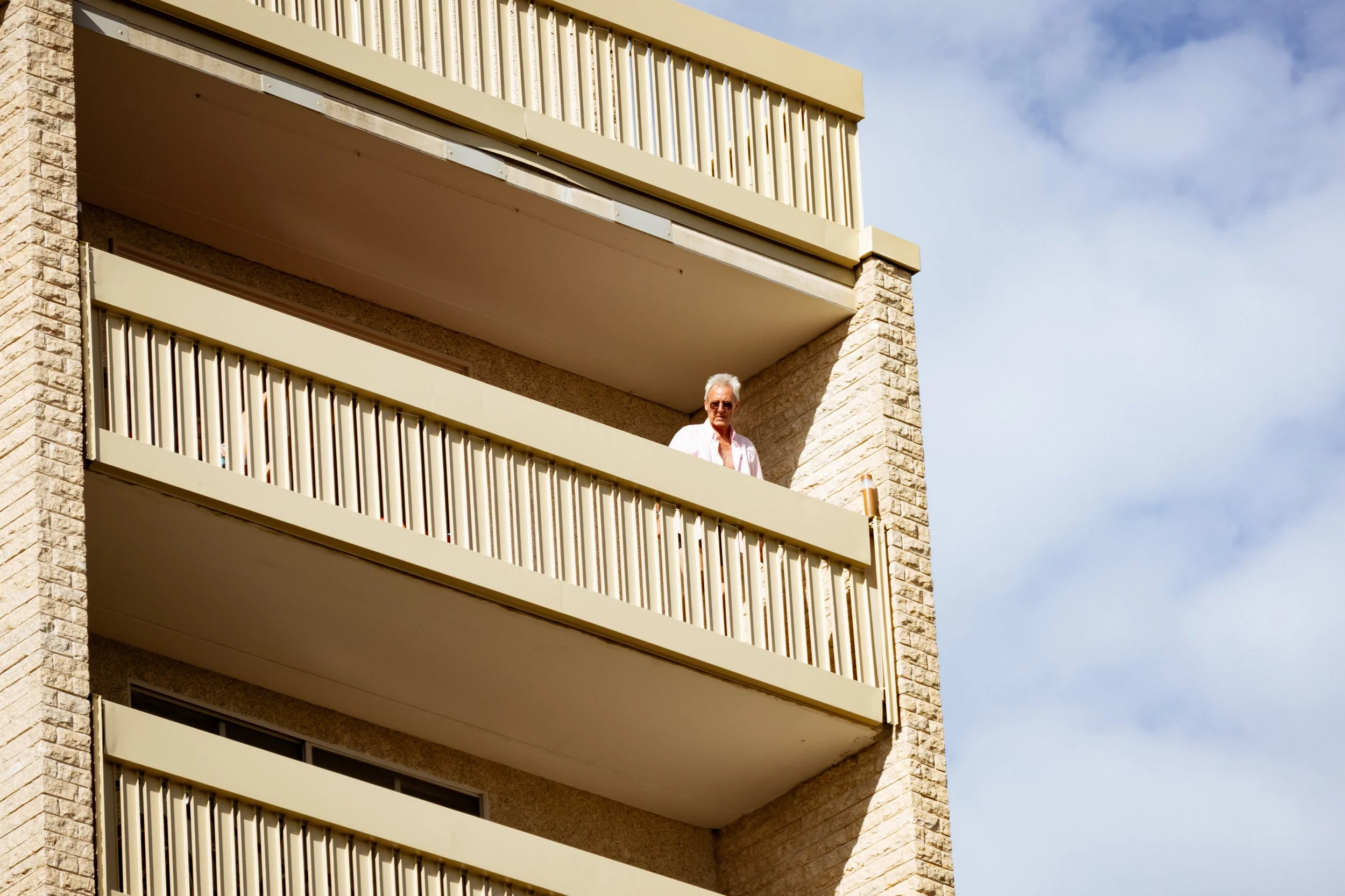 A man with white hair wearing sunglasses and a white shirt standing on a balcony of a modern apartment building made of brick and concrete, against a blue sky with some clouds.