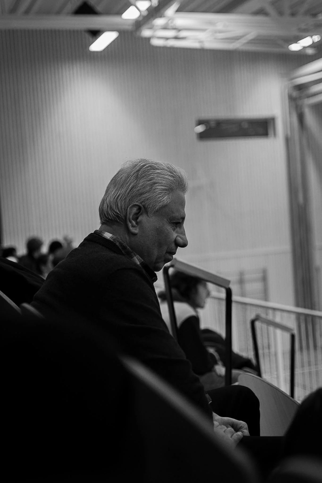 A black and white photo of an elderly man with gray hair sitting in a room, likely attending a meeting or conference, with other people seated behind him.