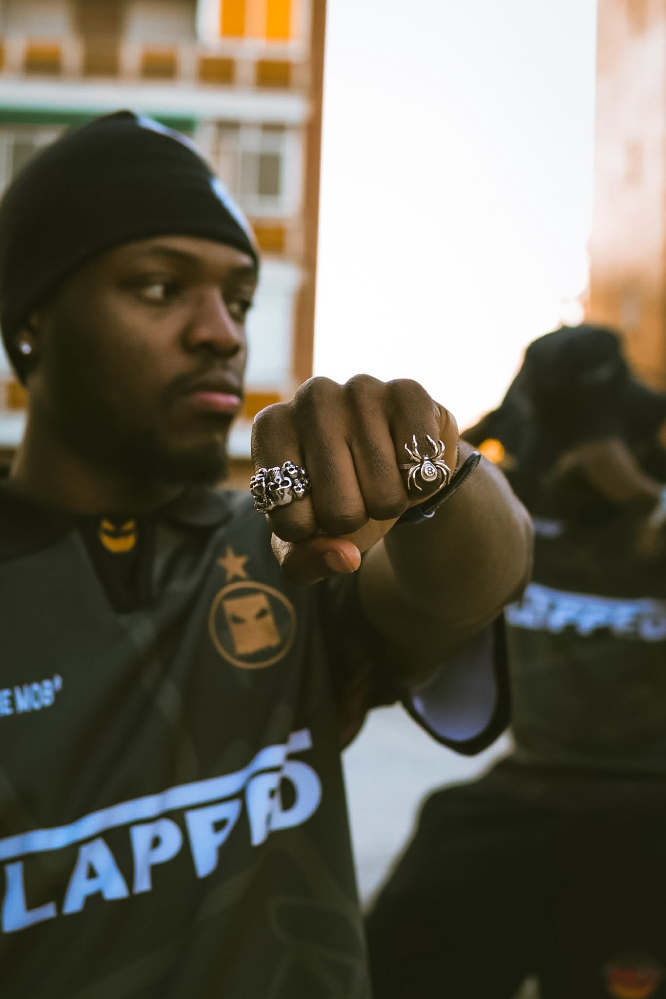 Close-up of a person wearing multiple rings, with one ring shaped like a spider. The person is dressed in black with a badge on their chest and is looking to the side, with part of their face visible.