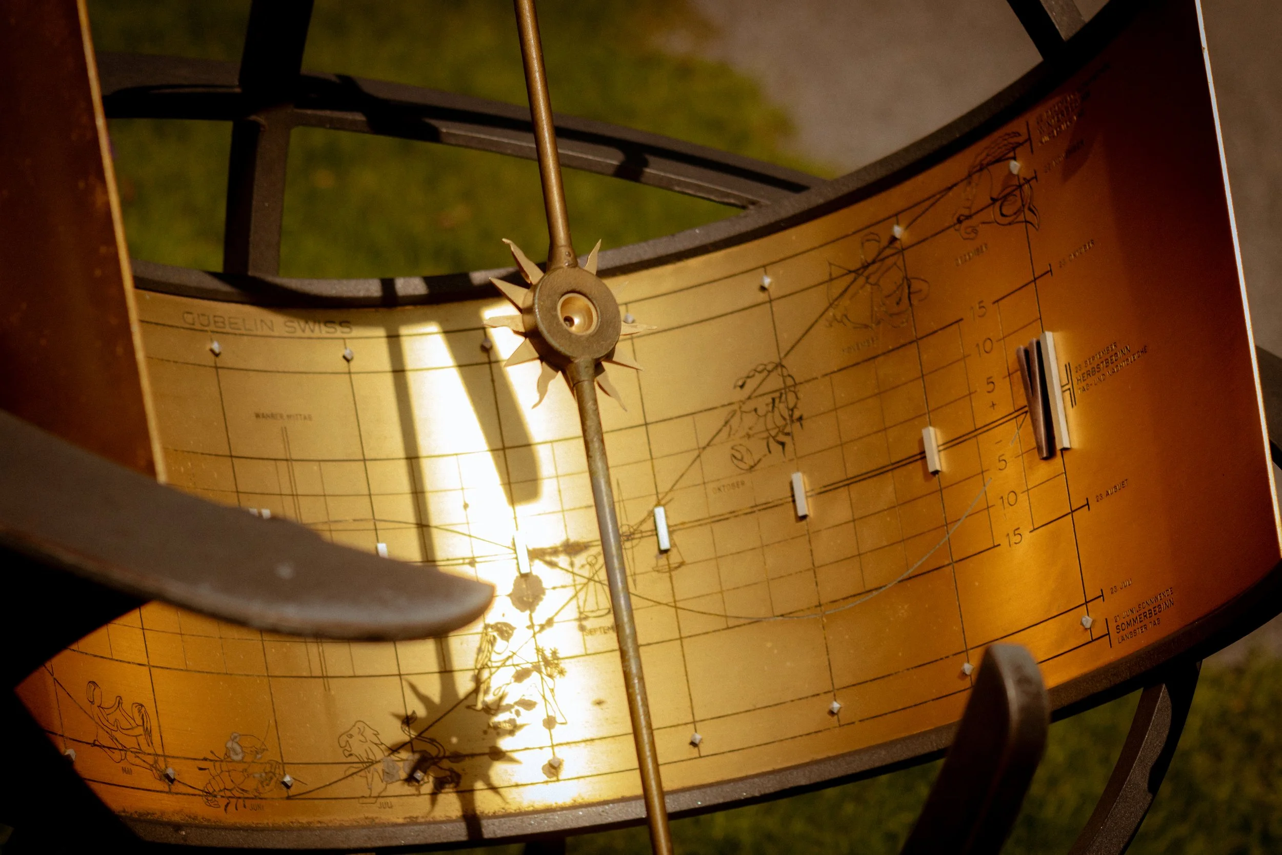 Close-up view of a vintage brass armillary sphere or celestial globe, mounted on a decorated wooden or metal stand, with a celestial and zodiac chart depicted on a golden background. The chart includes constellation illustrations and coordinate marki