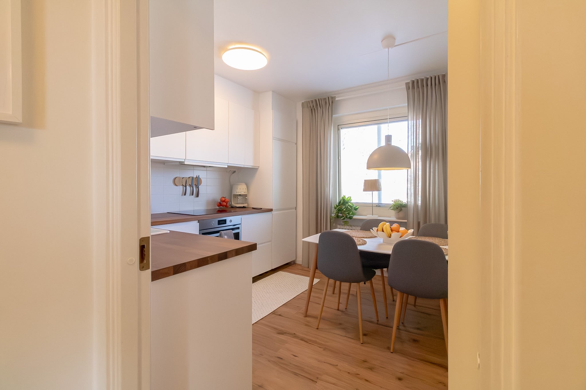 A view into a bright, modern kitchen and dining area with white cabinets, a window with beige curtains, a hanging white lamp, and a table with chairs and a bowl of fruit.