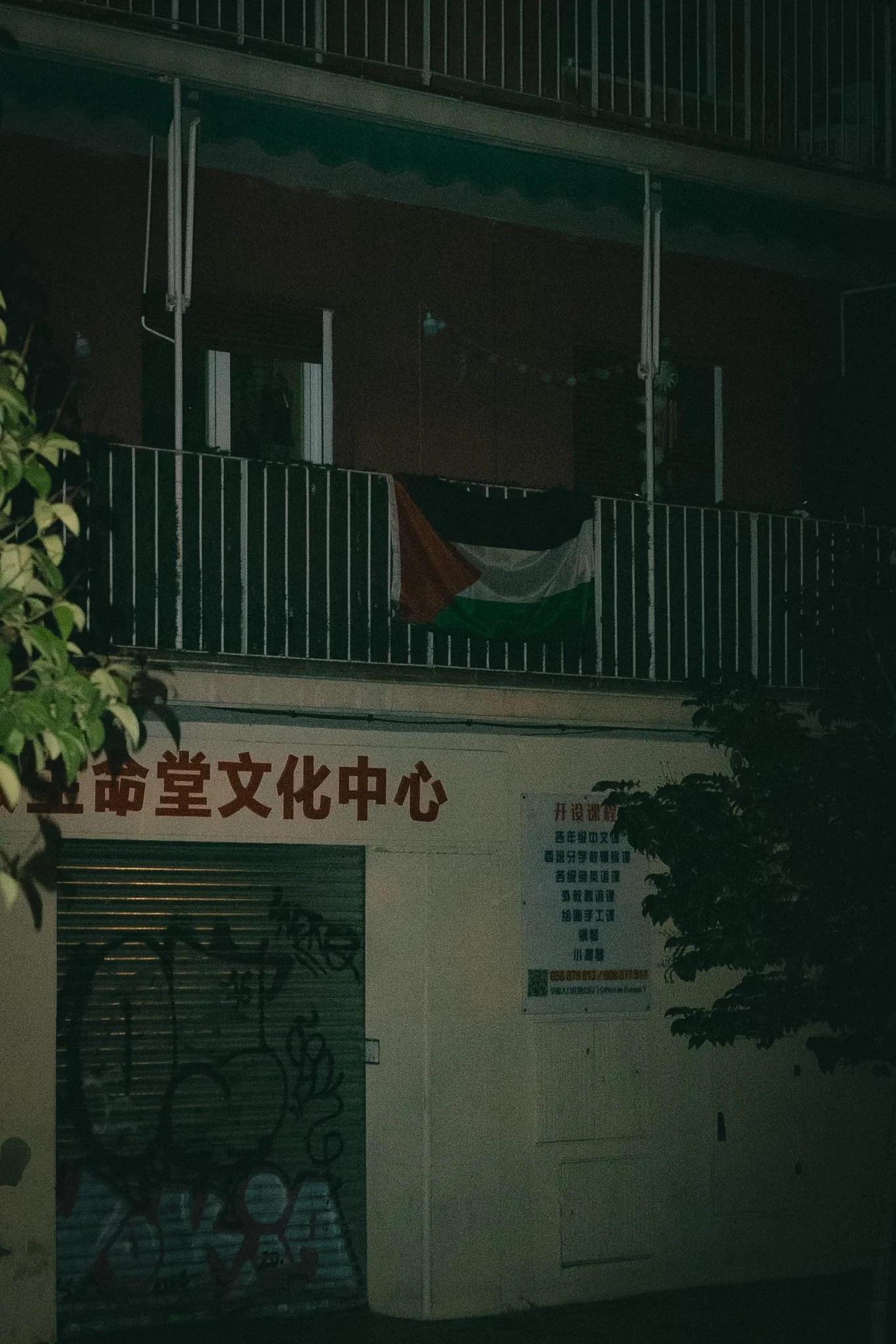 Nighttime view of a building with a sign in Chinese characters, a door, and a window. Some trees are visible in the foreground.