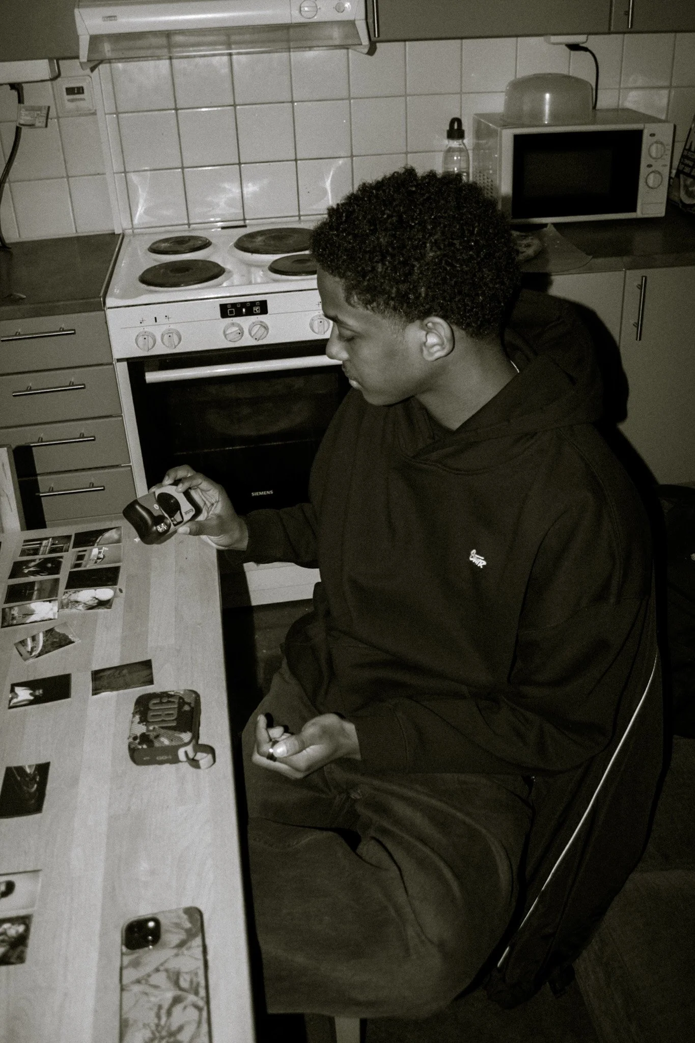 A young man with curly hair sitting at a kitchen table, holding a gaming controller, with photos and phone on the table, kitchen appliances including a stove, microwave, and bottles visible in the background.